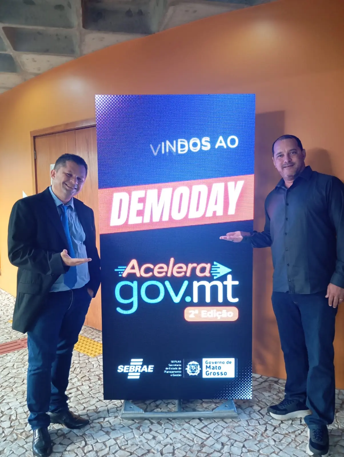 Dois homens, sorrindo, apontam para uma tela vertical que exibe "Vindos ao Demoday Acelera Gov.mt 2ª Edição" em cores vibrantes. Logos do Sebrae e Governo de MT no rodapé.