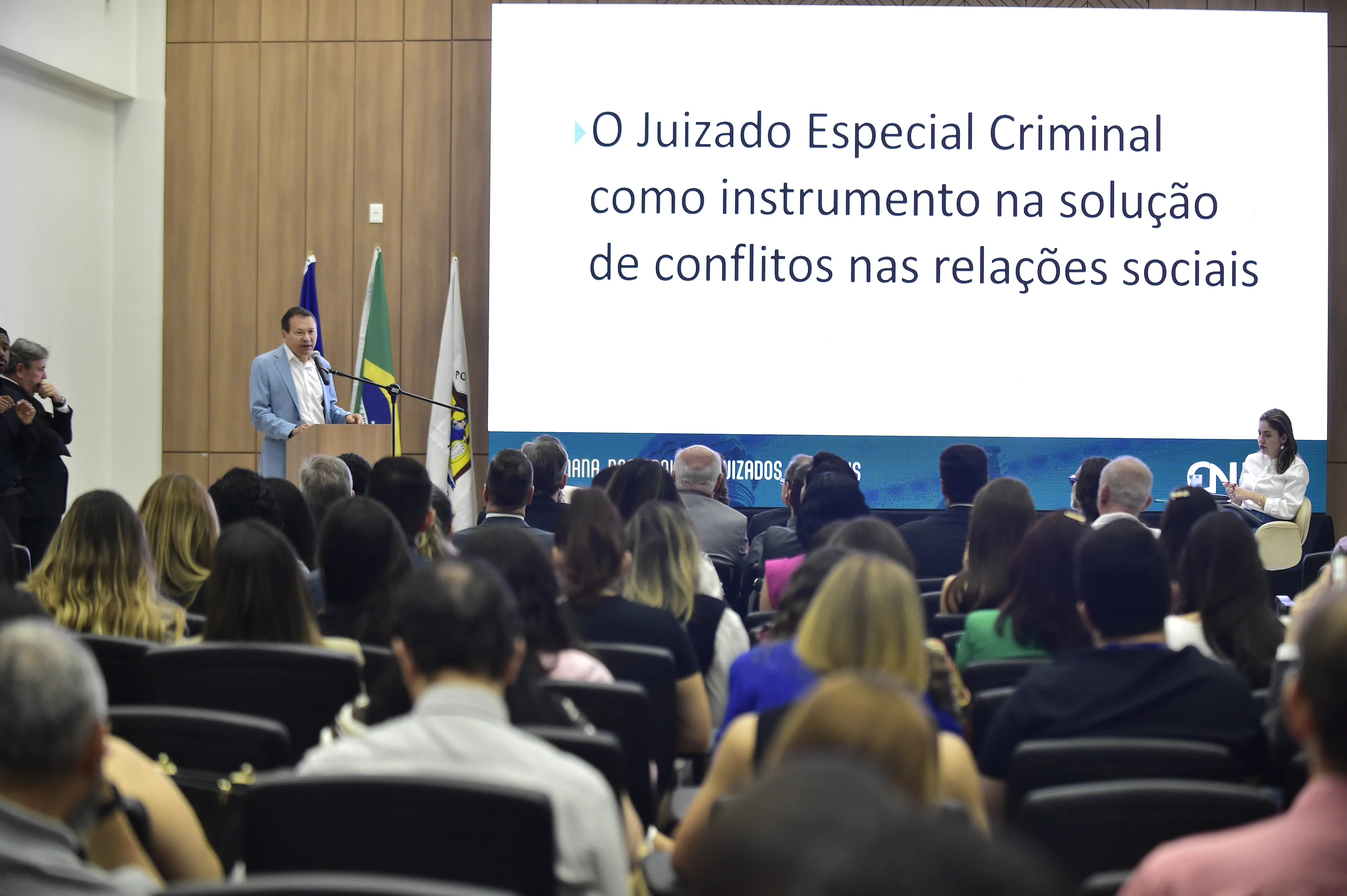 Em um auditório, um homem de terno claro discursa em púlpito. Atrás dele, bandeiras e um telão branco exibindo o tema 'O Juizado Especial Criminal como instrumento na solução de conflitos nas relações sociais'. A plateia o observa