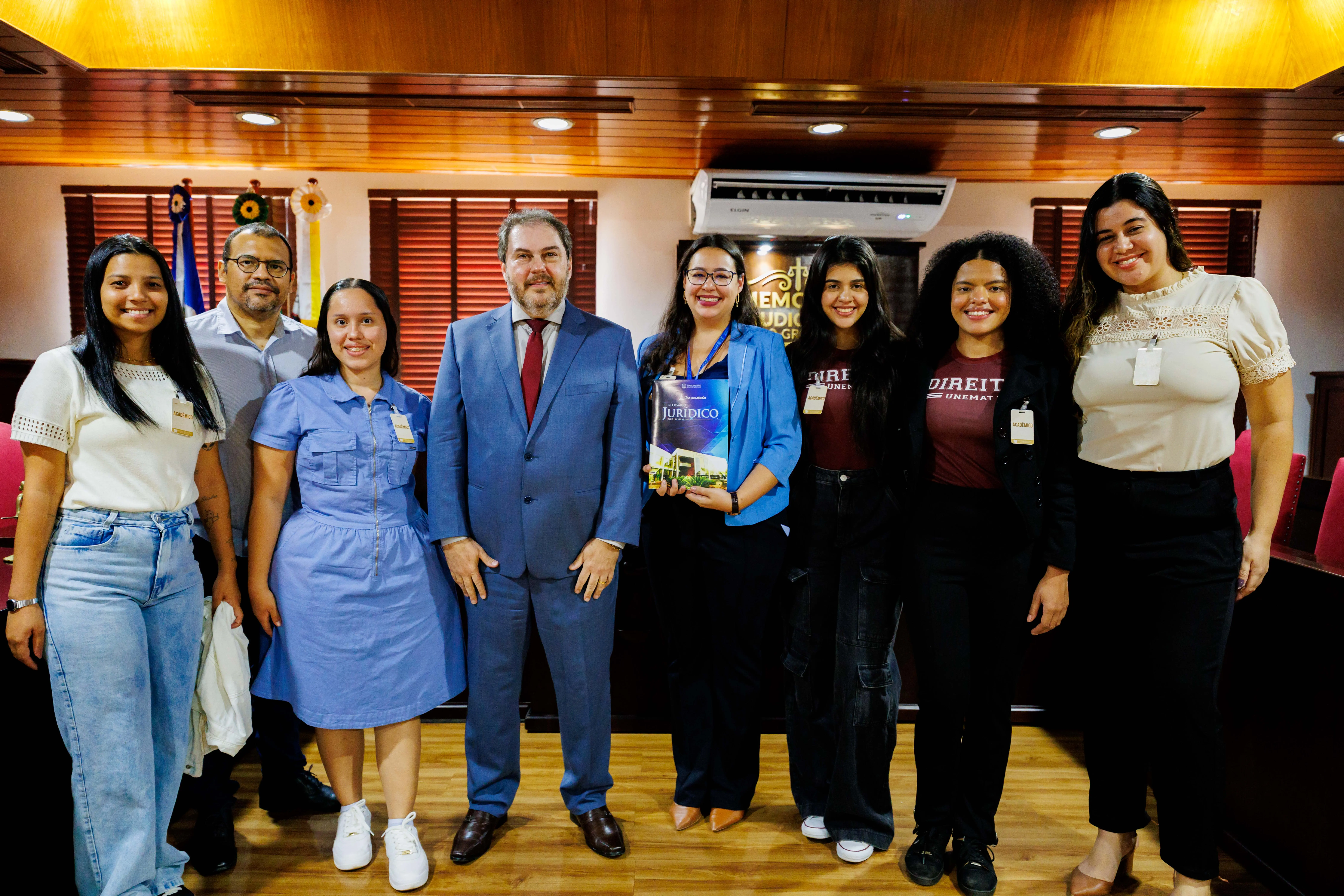 Grupo de oito pessoas posa sorridente no Memorial do Judiciário Mato-grossense. Ao centro, homem de terno azul e gravata vermelha ladeado por acadêmicos, alguns com camisetas de Direito da Unemat e crachás.