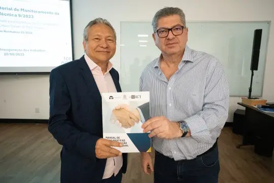 Dois homens posam sorrindo para foto segurando o “Manual de Boas Práticas da Governança Social”, durante encontro no Tribunal de Contas de Mato Grosso.