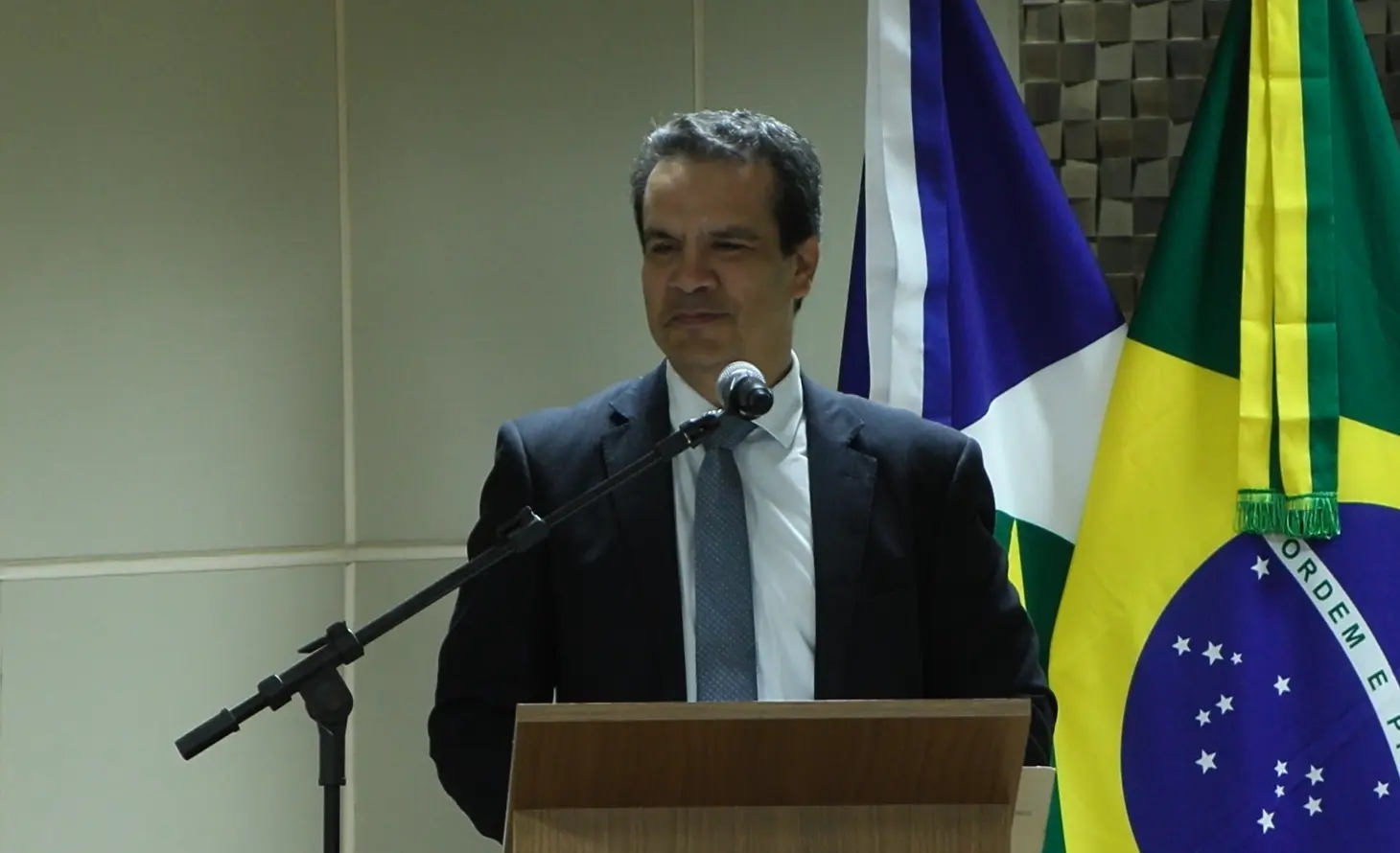 Juiz Emerson Cajango falando no púlpito do auditório do TJ. Ele é um homem pardo, de cabelos e olhos escuros, usando camisa clara, gravata cinza e terno azul marinho. Atrás deles estão as bandeiras do Brasil e de Mato Grosso. 