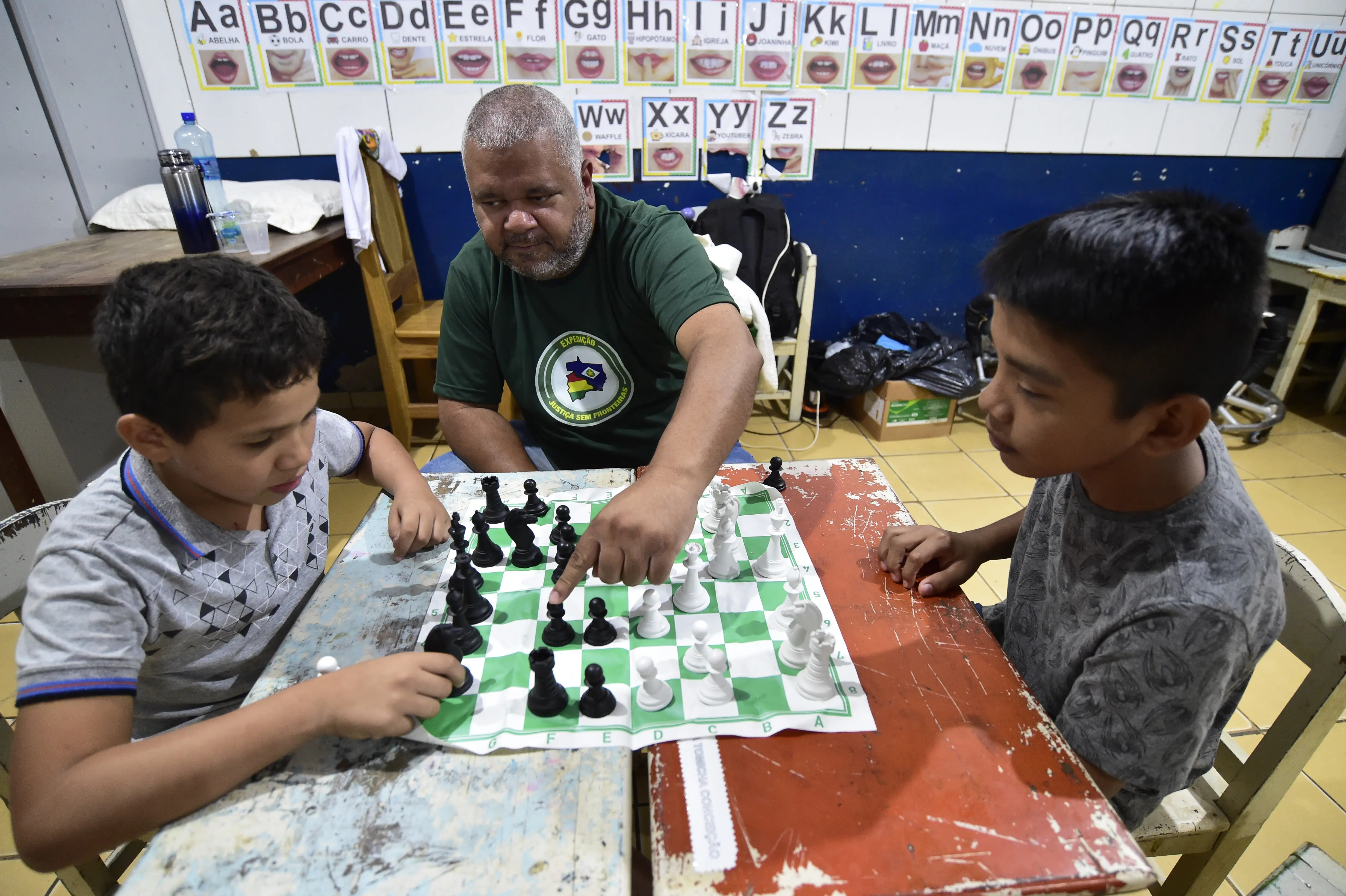 Dois meninos aprendem a jogar xadrez com auxílio de um servidor da Secretaria de Esporte, que está sentado ao lado da mesa, entre eles. Eles estão em uma sala de aula.