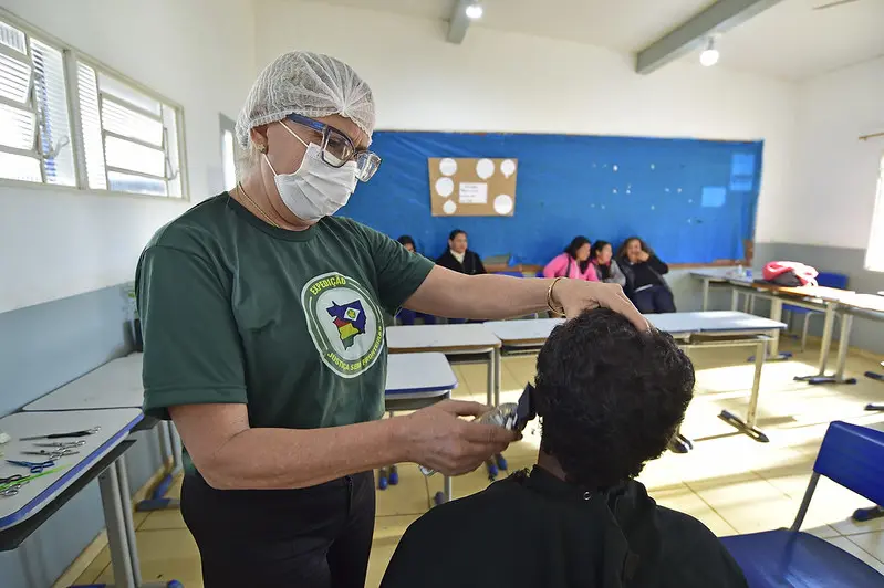 Mulher usando camiseta da Justiça Sem Fronteiras, máscara e touca descartáveis corta o cabelo de um homem na máquina, em uma sala de aula.