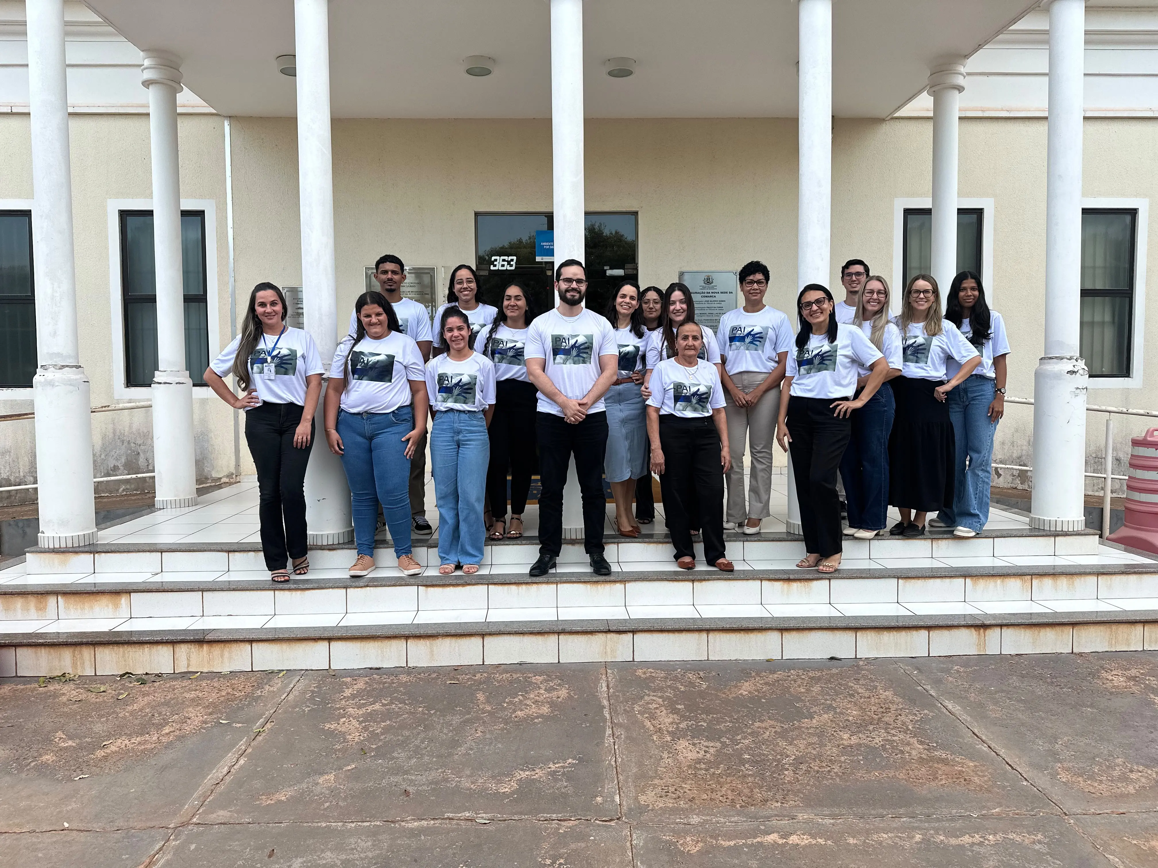 Grupo de pessoas posa nas escadas de um prédio com colunas brancas. Todas vestem camisetas brancas padronizadas da campanha "Pai Presente" e calças escuras ou jeans.