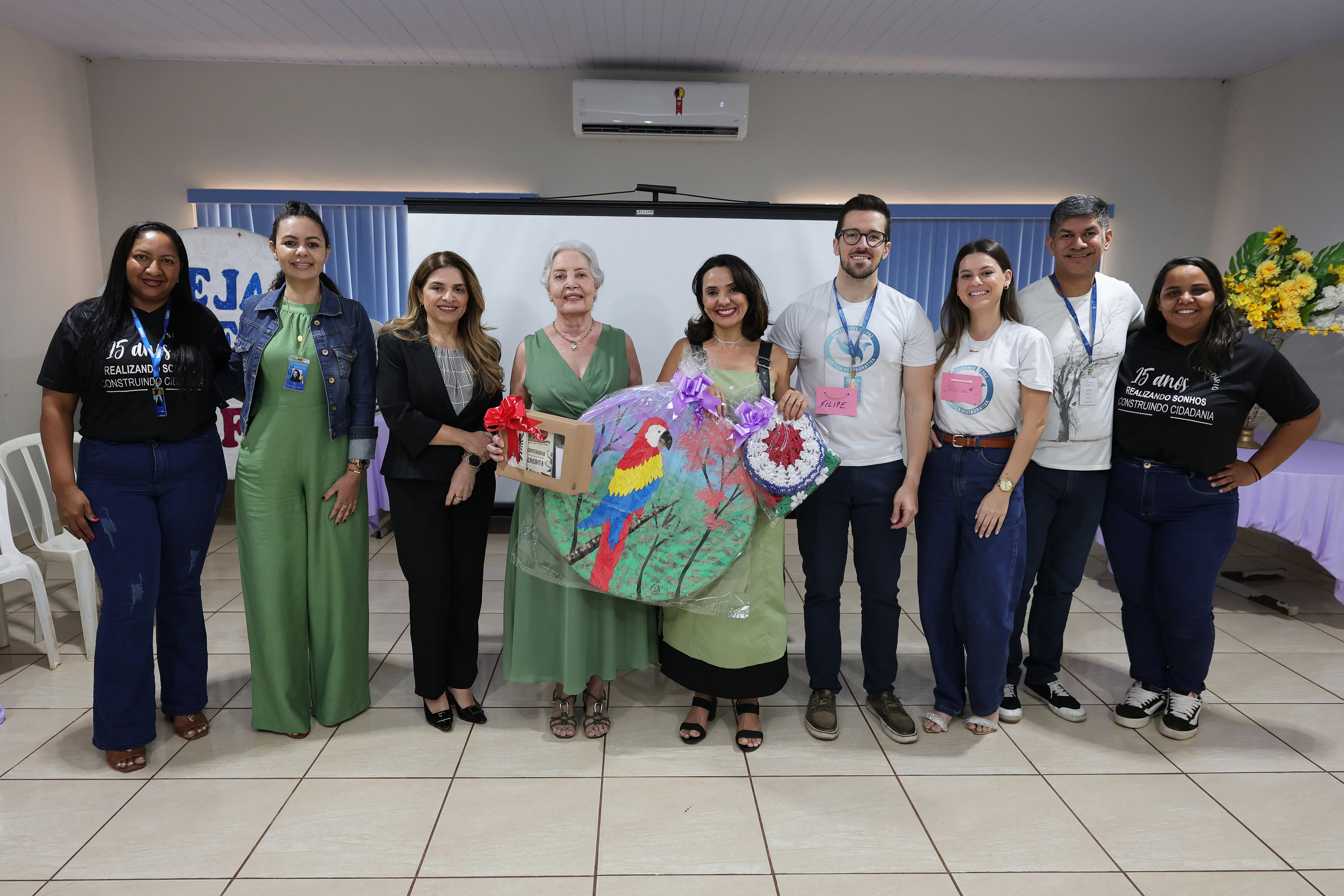 Grupo de nove pessoas sorri alinhado em uma sala. Ao centro, duas mulheres seguram presentes, incluindo uma tela redonda com pintura de uma arara colorida e uma caixa decorada com laço vermelho.