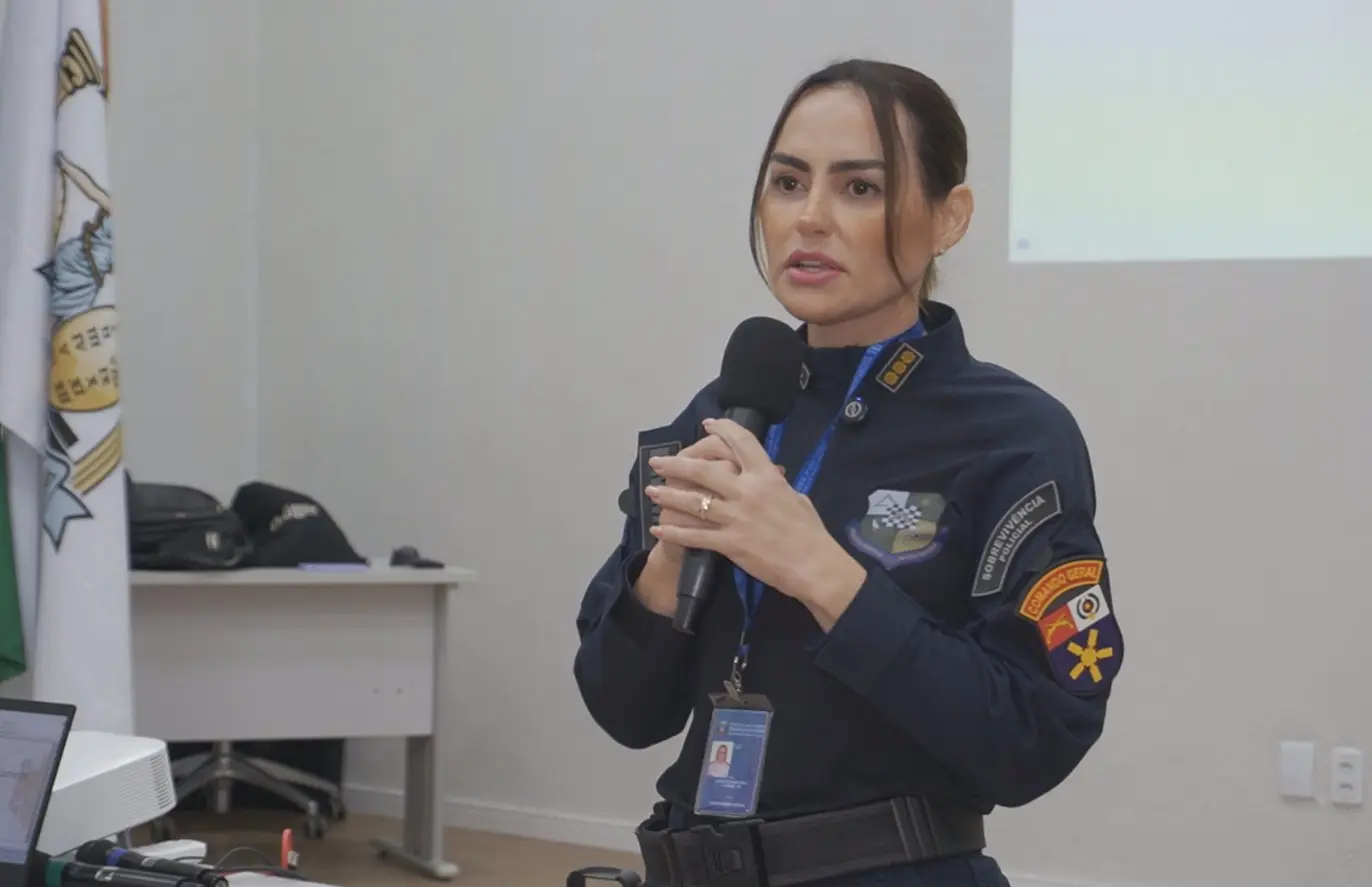 Foto horizontal em plano médio que mostra a coordenadora militar do TJMT, coronel PM Jane, em pé, falando ao microfone. Ela é uma mulher parda, de cabelos castanhos e presos, usando farda azul marinho da Polícia Militar.