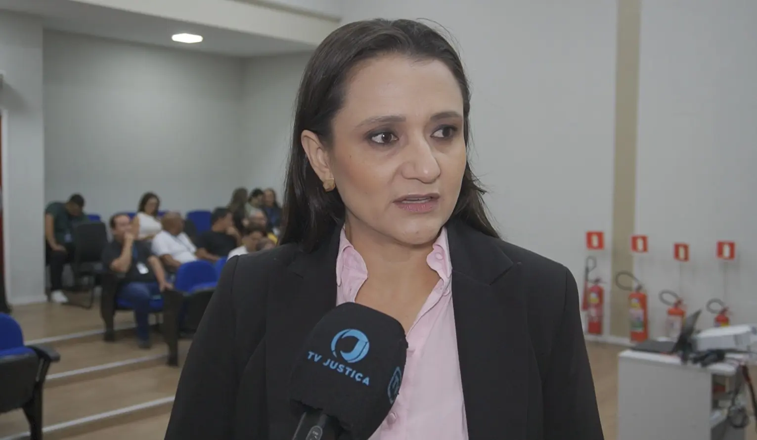 Foto horizontal que mostra a chefe da Divisão de Prevenção e Combate à Incêndio do TJMT, tenente-coronel Bombeiro Aline Novacki, durante entrevista à TV Justiça. Ela é uma mulher de pele clara, olhos e cabelos castanhos, usando camisa rosa clara e blazer preto. Atrás dela, há pessoas sentadas. 