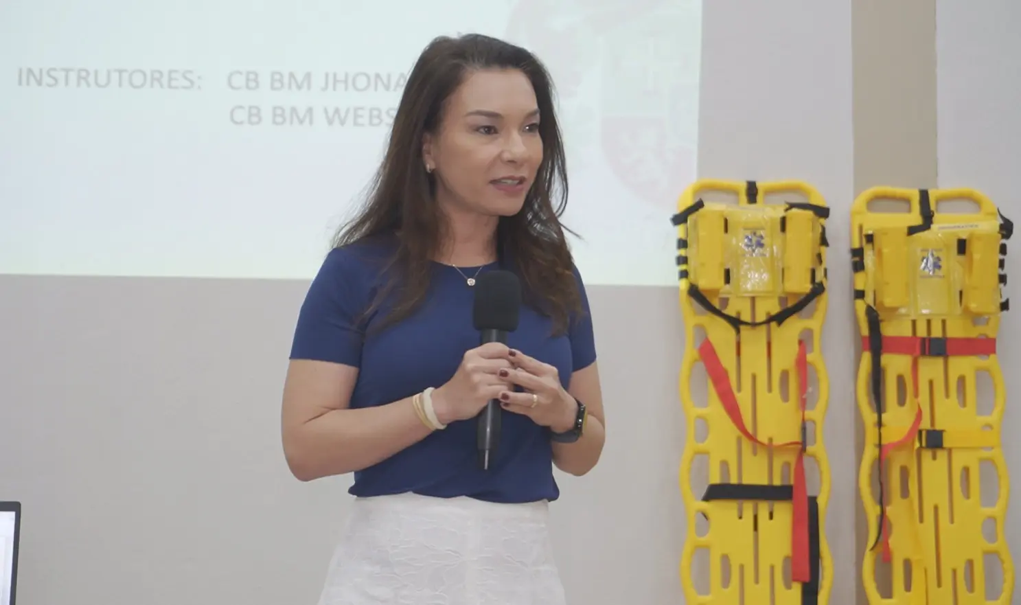 Foto horizontal que mostra a juíza Hannae Yamamura em pé, falando ao microfone, no auditório do Fórum de Cuiabá, na abertura do curso de brigadista. ela é uma mulher branca, de traços orientais, cabelos longos e castanhos, usando saia branca e blusa azul marinho.