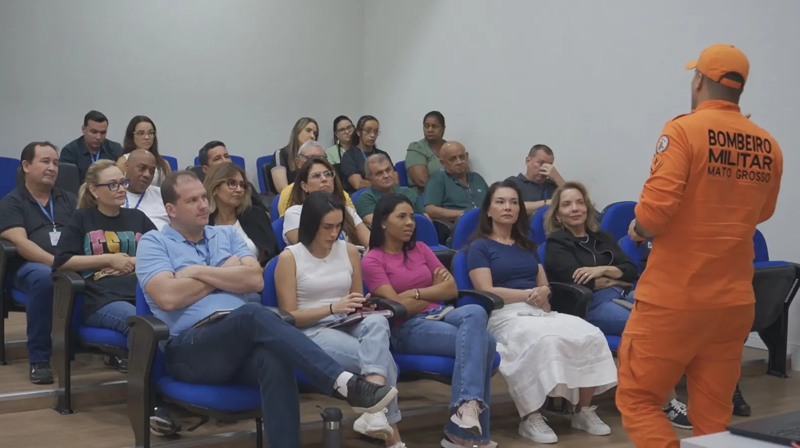 foto horizontal em plano aberto que mostra cerca de 20 pessoas sentadas, assistindo a uma palestra de um bombeiro militar, que está em pé, com farda laranjada. 