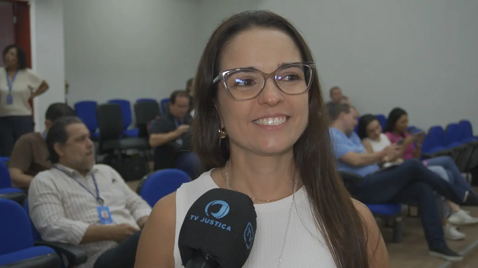 Foto horizontal que mostra a servidora Laura Martine durante entrevista à TV Justiça. Ela é uma mulher de pele clara, cabelos longos, lisos e castanhos, usando óculos de grau de armação cinza e blusa regata branca. Atrás dela, há várias pessoas sentadas no auditório. Laura sorri.