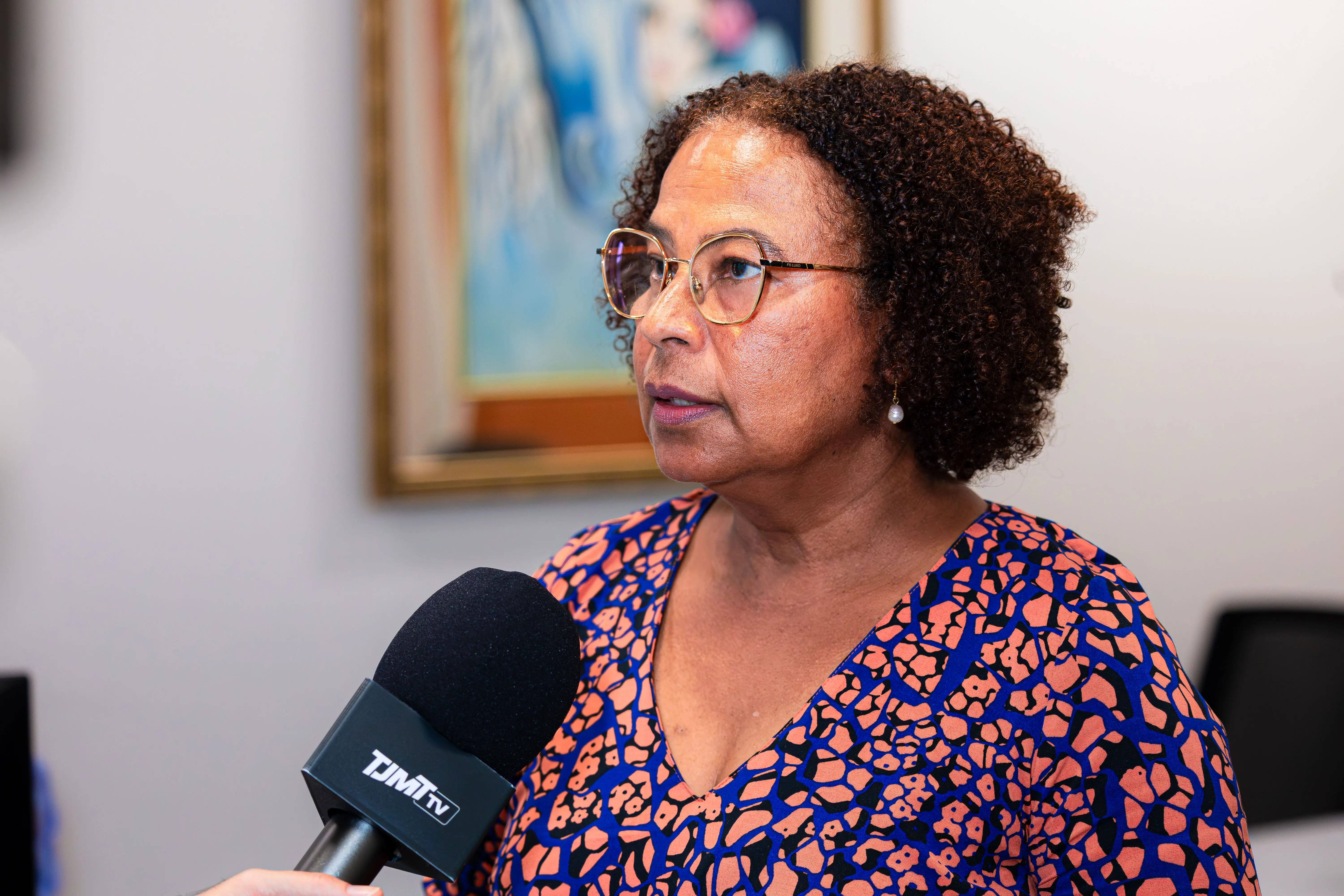 Mulher de cabelos cacheados escuros e óculos fala ao microfone com a marca "TJMT TV". Veste blusa estampada em tons de azul e laranja. Ao fundo, quadro emoldurado.