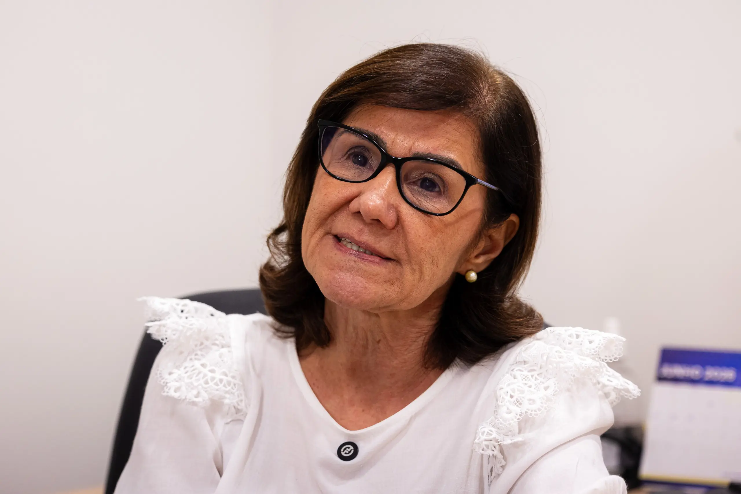 Mulher de cabelos castanhos curtos e óculos de armação escura olha ligeiramente para cima. Usa blusa branca com detalhes em renda nos ombros. Ao fundo, parede e calendário desfocado.