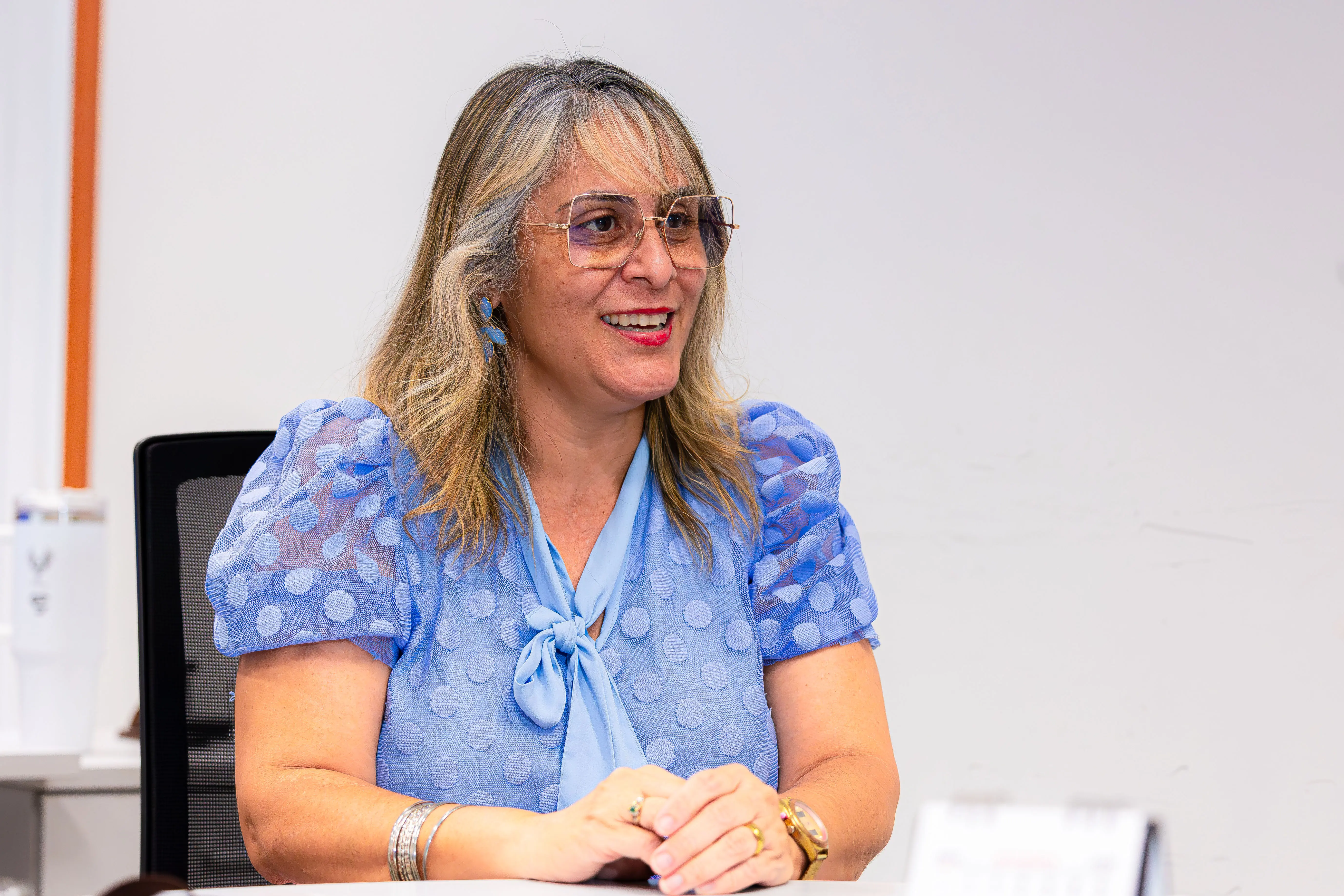 Mulher sorridente de óculos de armação clara e cabelos claros com mechas veste blusa azul texturizada de mangas curtas bufantes. Está sentada à mesa com as mãos apoiadas.