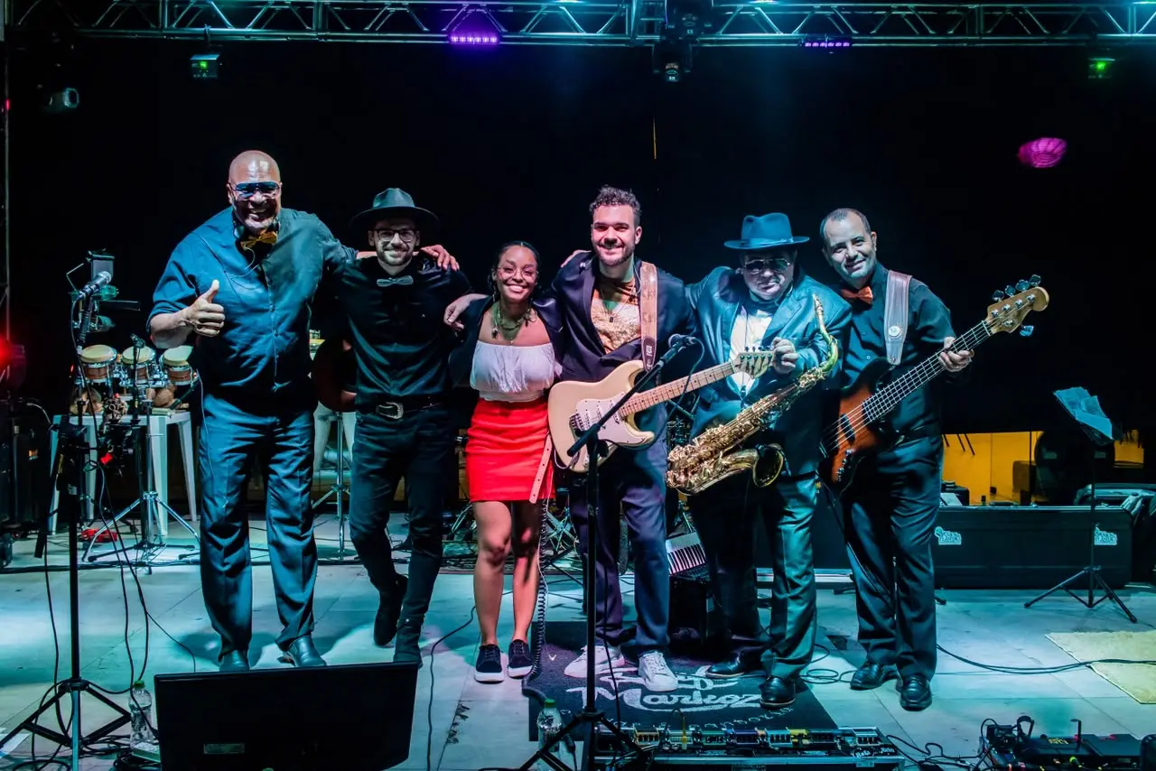 Seis integrantes da banda posam abraçados no palco: mulher de saia vermelha ao centro, dois homens de chapéu, um com saxofone, outro com guitarra e outro com baixo.