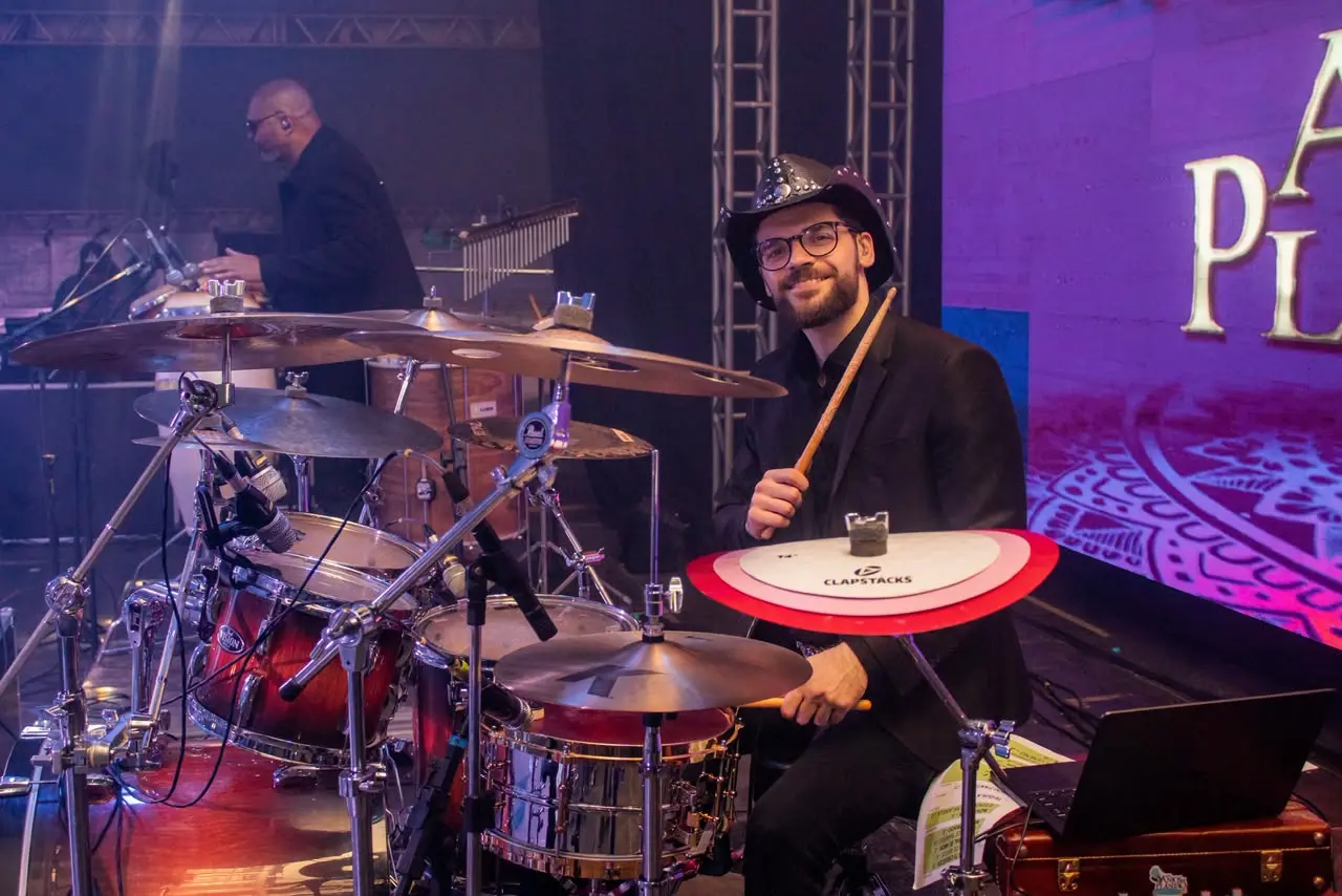 Homem de barba, óculos e chapéu escuro com brilhos sorri e segura baquetas, sentado à bateria. Veste terno preto e, ao fundo, outro músico toca sob luzes roxas.