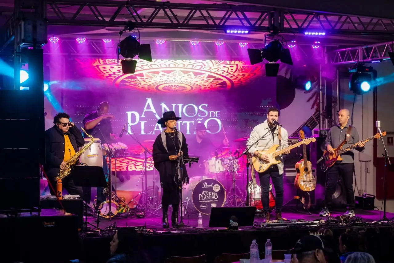 Banda Anjos de Plantão se apresenta no palco: mulher de chapéu ao microfone, saxofonista, guitarrista, baixista e baterista, sob luzes roxas e painel com o nome do grupo.