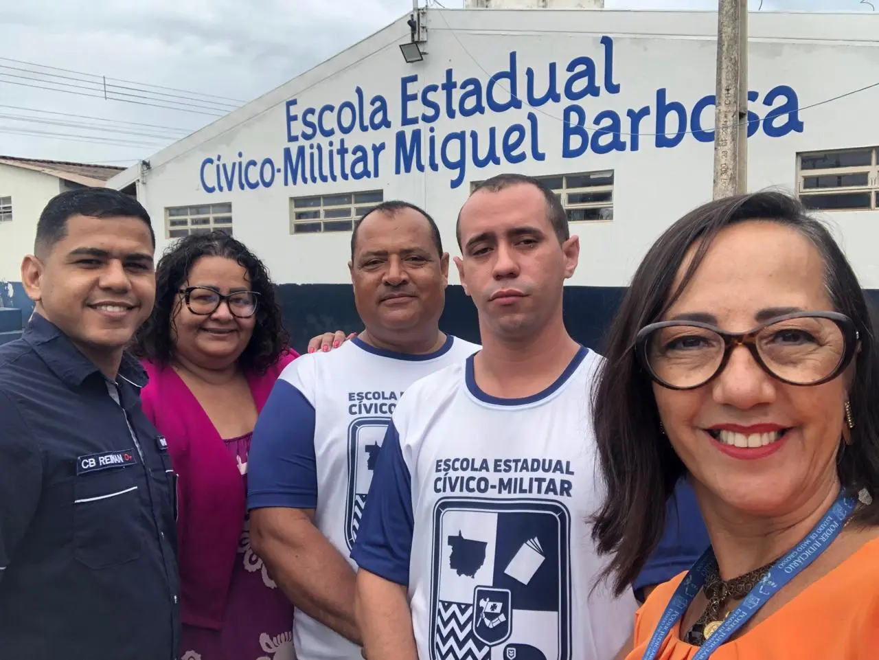 Foto que mostra a agente da infância e juventude de São José dos Quatro Marcos ao lado de outros servidores públicos e profissionais da Escola Miguel Bandeira posando para a foto em frente à fachada da unidade escolar. 