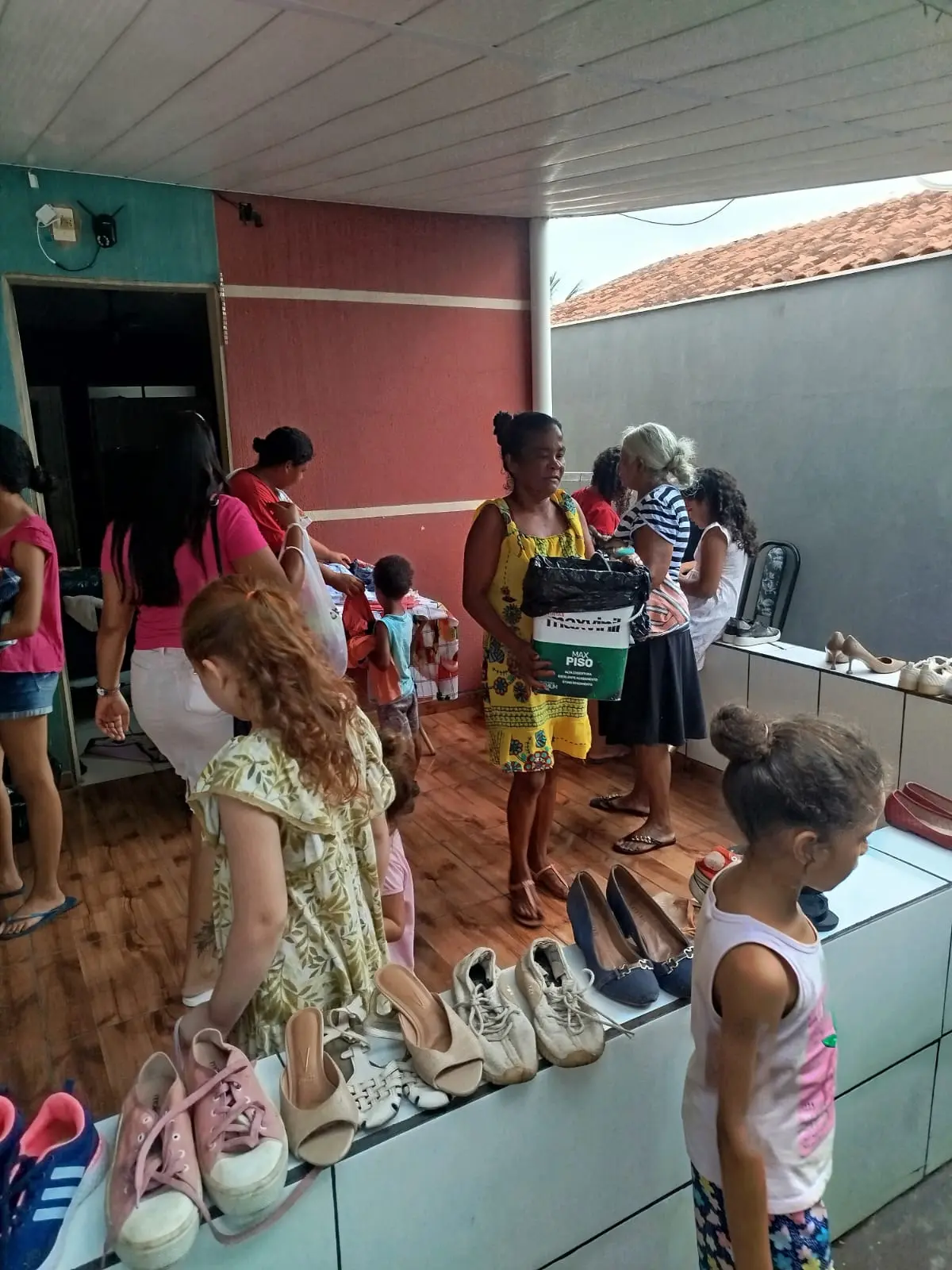 Grupo de mulheres e crianças reúne-se em espaço comunitário para escolher roupas sobre mesa azul. A ação demonstra cuidado coletivo, organização e apoio social.