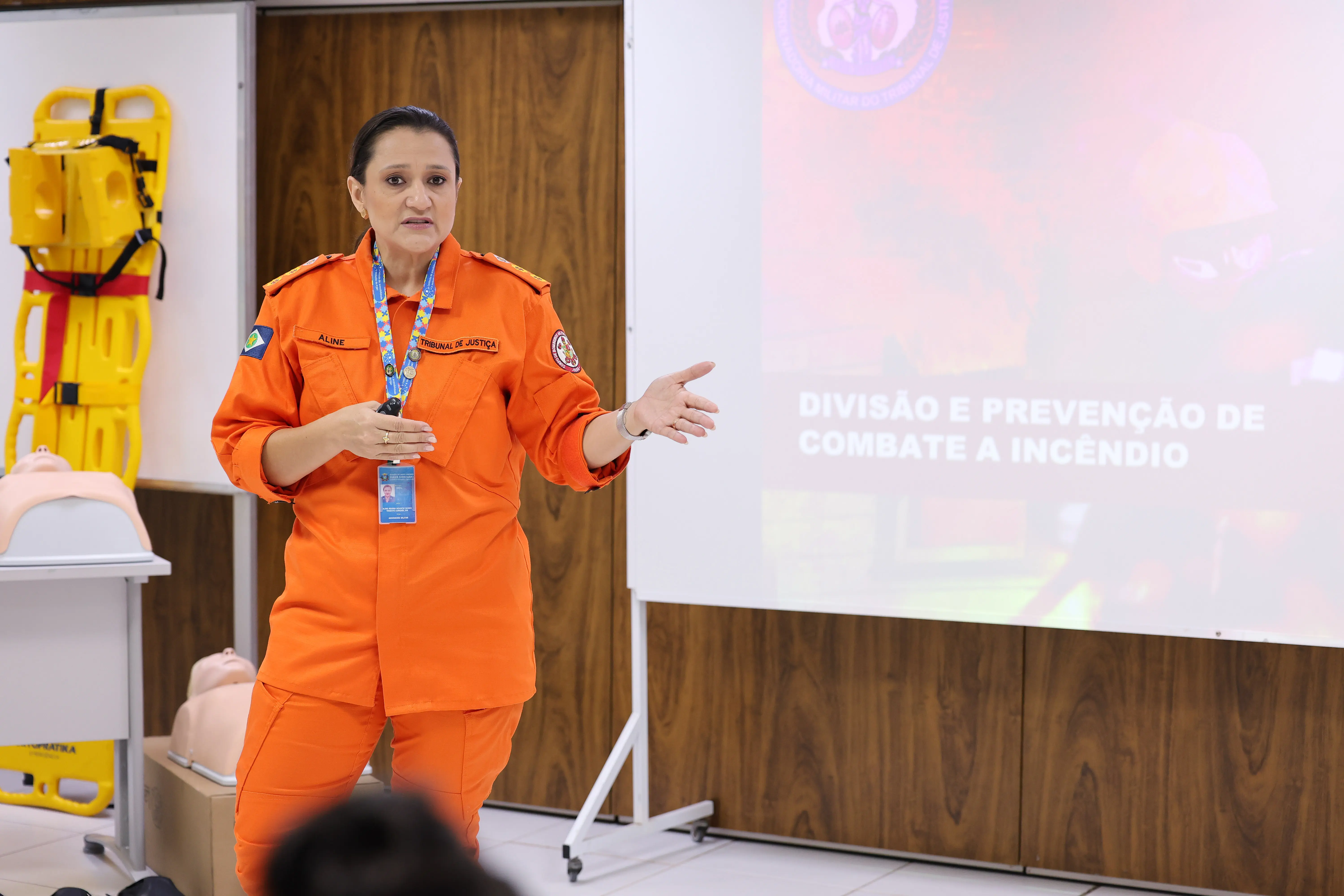 Uma bombeira fardada de laranja faz uma apresentação. Ela gesticula com as mãos ao lado de um telão com o texto 