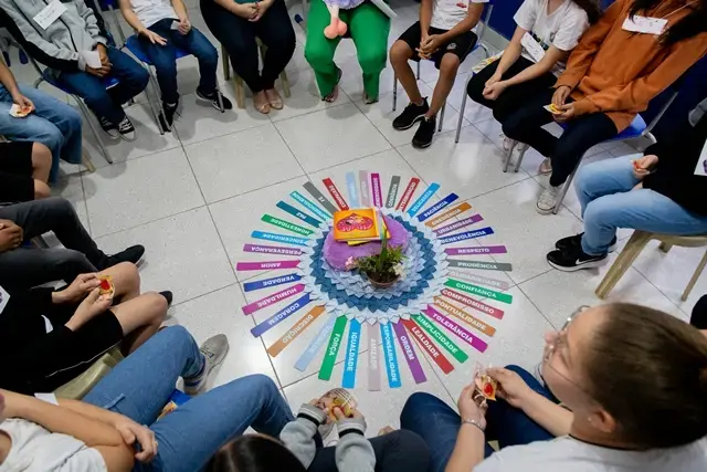 Grupo de pessoas sentadas em círculo dentro de uma sala participa de atividade coletiva. No centro, há cartazes coloridos com palavras e um arranjo simbólico. O ambiente é fechado, com piso claro, e a dinâmica sugere escuta, diálogo e fortalecimento de vínculos.