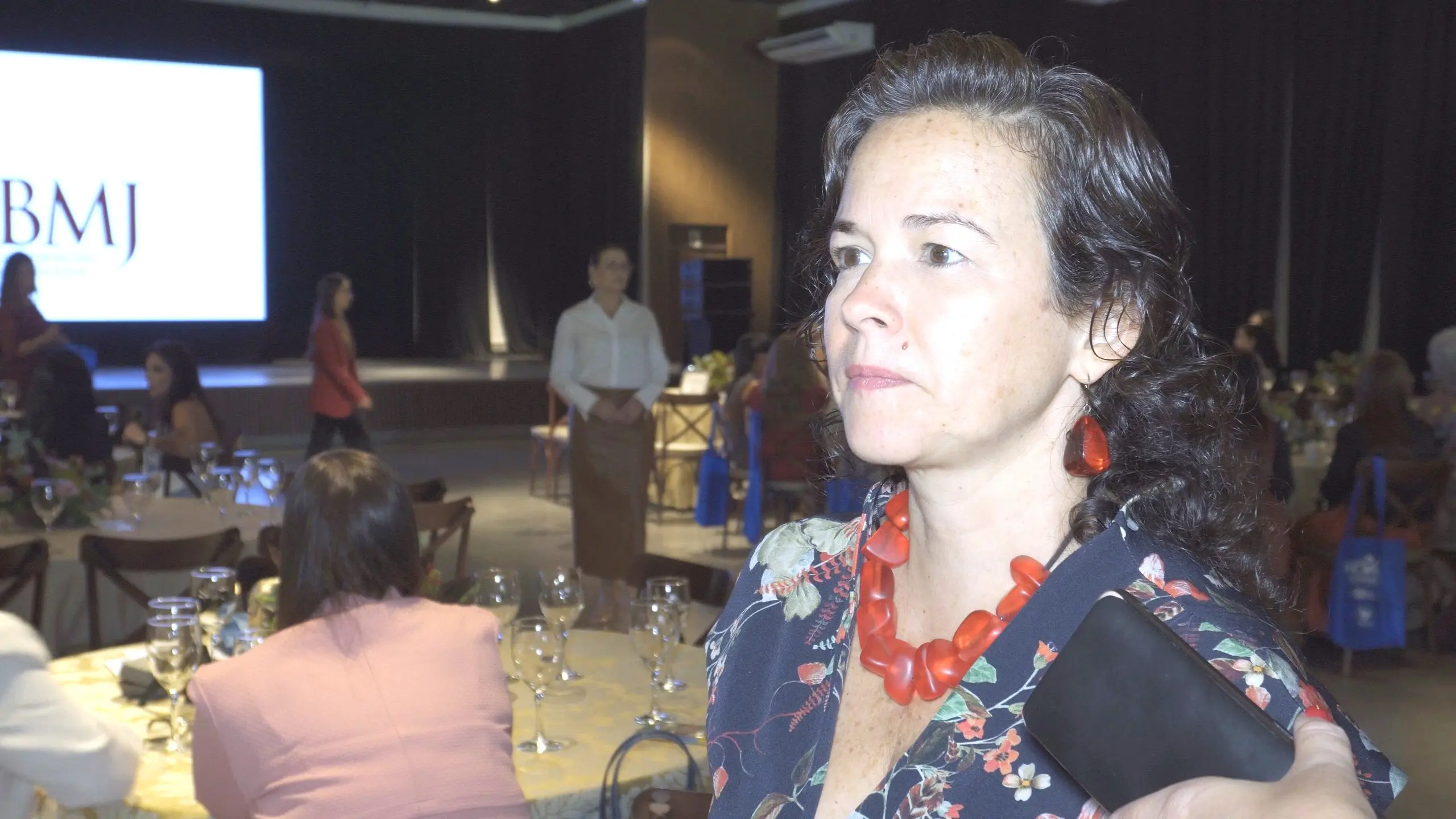 Uma mulher de meia-idade, com cabelos escuros, veste um vestido preto justo com estampa floral e sapatos vermelhos de salto alto. Ela está em pé, em um palco, segurando dois microfones e falando. Um telão branco com a imagem de um homem de terno azul está atrás dela.