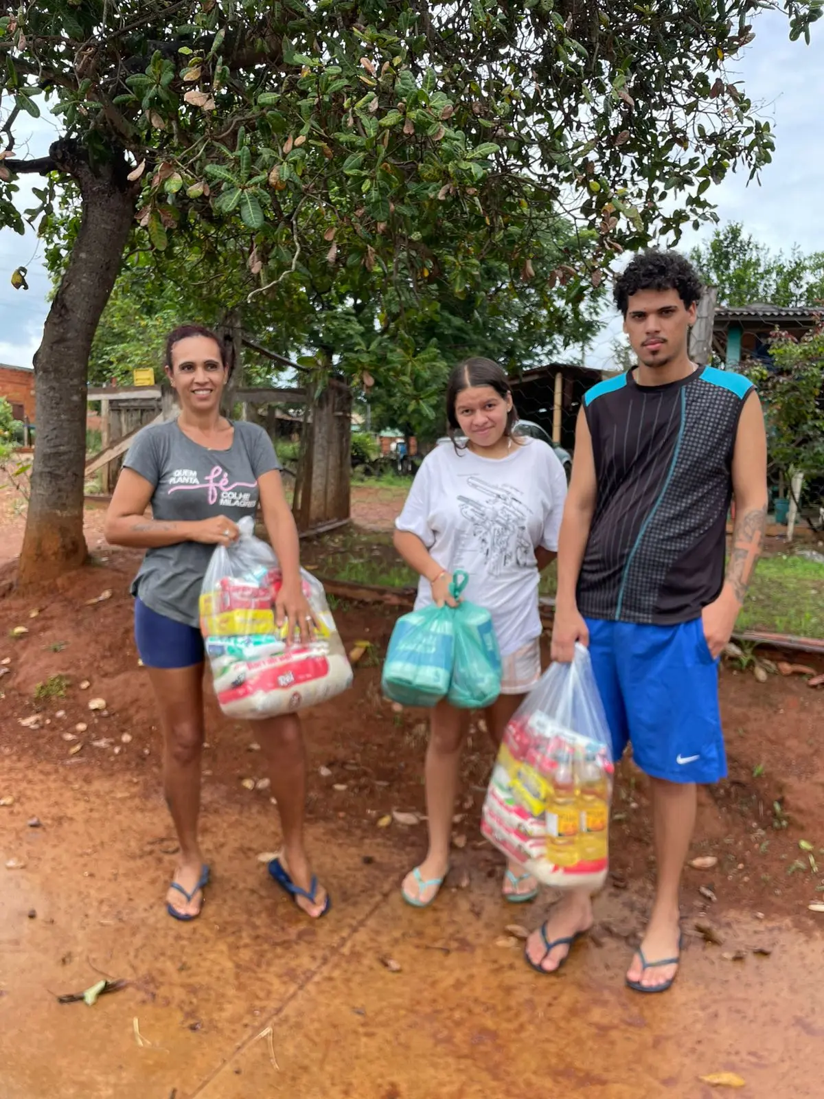 Três pessoas posam com cestas de alimentos nas mãos, em quintal arborizado. A imagem simboliza solidariedade, apoio comunitário e fortalecimento de vínculos locais.