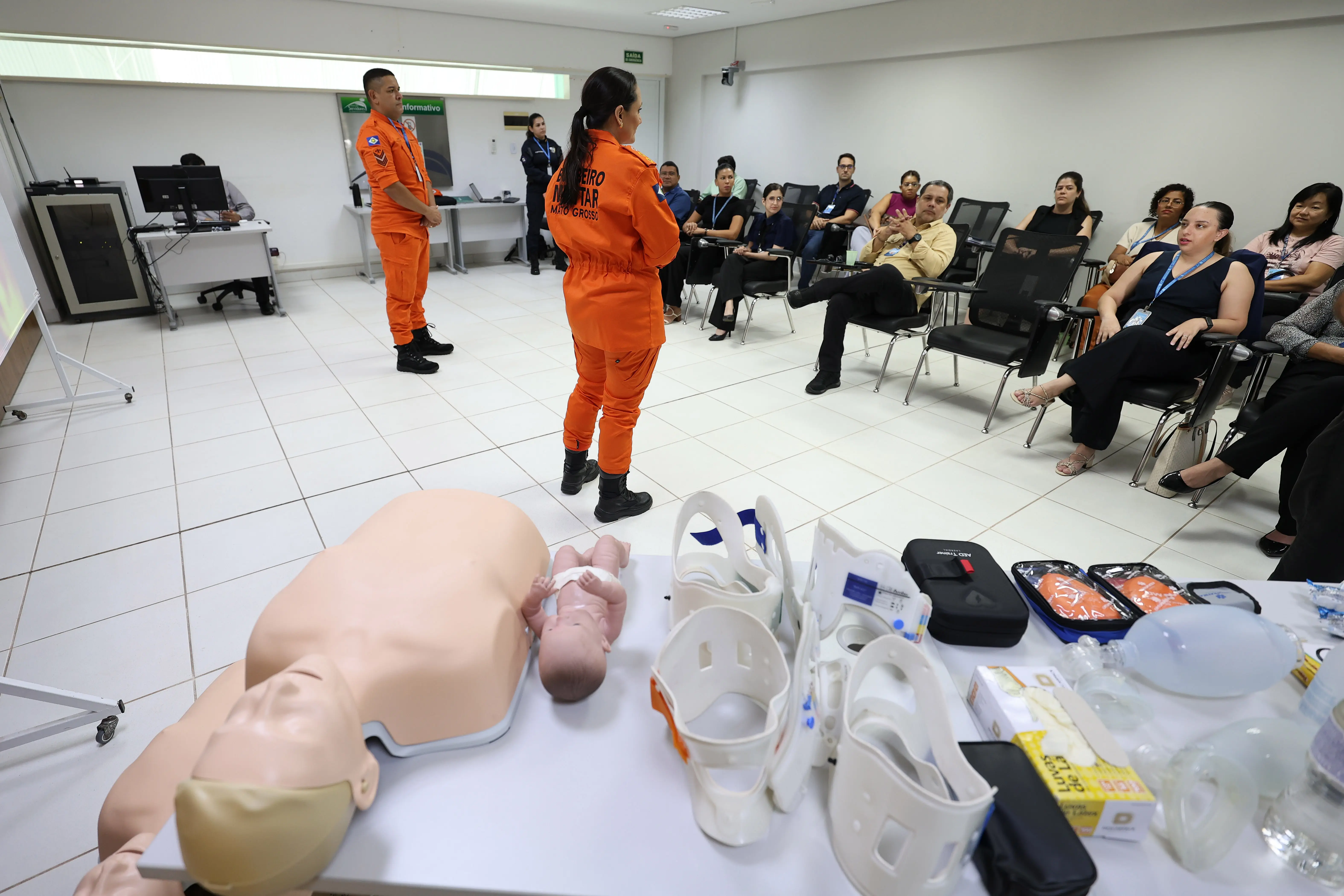 Uma sala de treinamento com bombeiros fardados de laranja ensinando primeiros socorros a um grupo sentado. Em primeiro plano, uma mesa com manequins de simulação (adulto e bebê), colares cervicais e equipamentos médicos. O ambiente é claro e profissional.