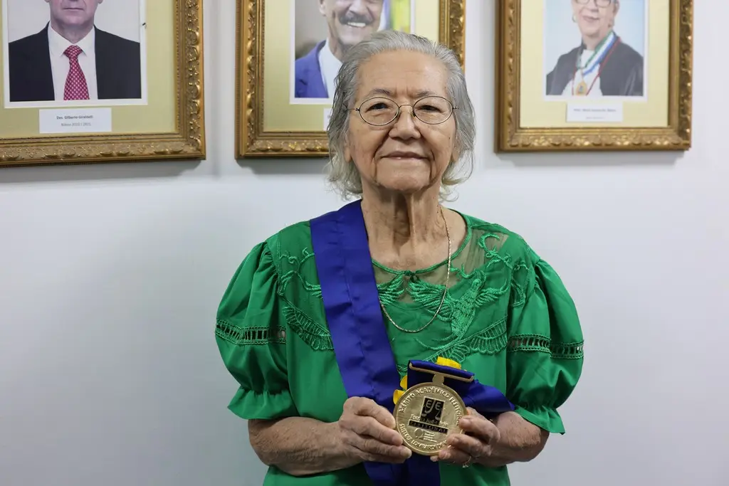 Desembargadora aposentada Shelma Lombardi de Kato posa sorrindo para a foto, segurando a medalha do mérito acadêmico da Escola Judiciária Eleitoral, que leva seu nome. Ela é uma idosa, com traços orientais, cabelos lisos e brancos, usando blusa verde e uma faixa azul com a medalha.