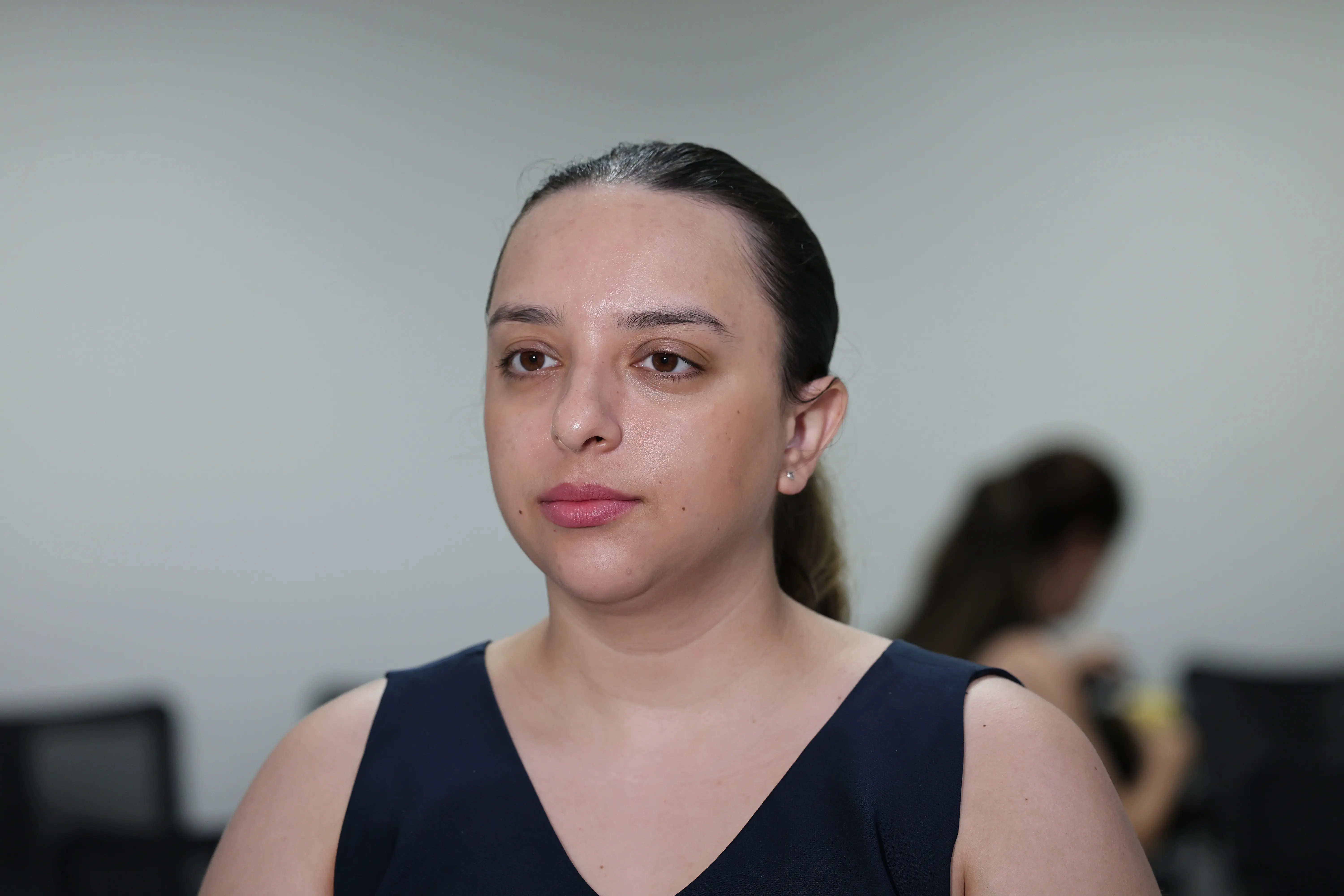 Mulher jovem de pele clara e cabelos escuros presos, vestindo uma regata azul-marinho. Ela tem uma expressão serena e atenta, olhando levemente para o lado. O fundo é uma parede clara e neutra, com pessoas desfocadas ao fundo