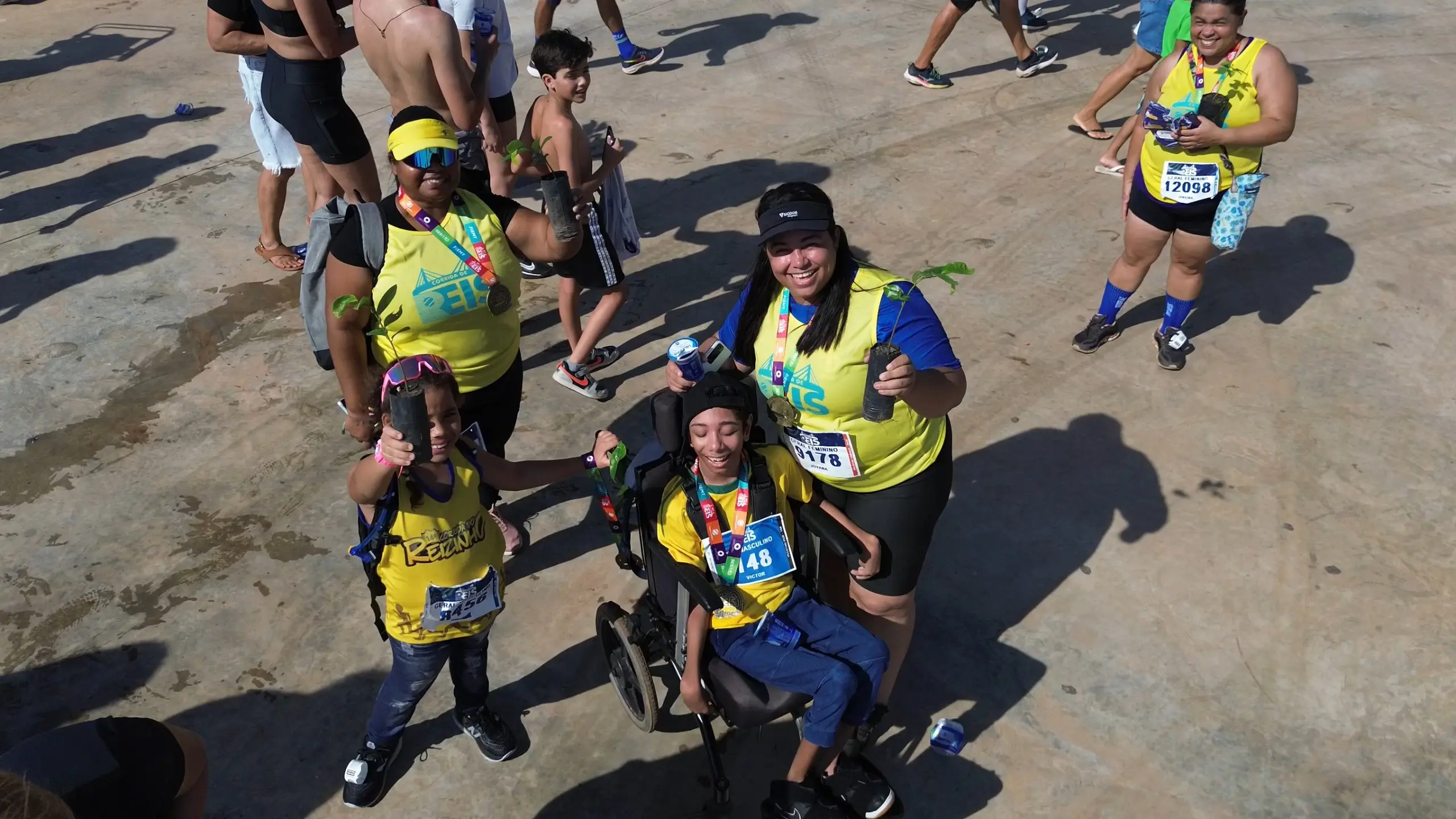 Grupo de corredores com medalhas e camisas amarelas, incluindo uma pessoa em cadeira de rodas, celebra segurando mudas de árvores após a corrida.