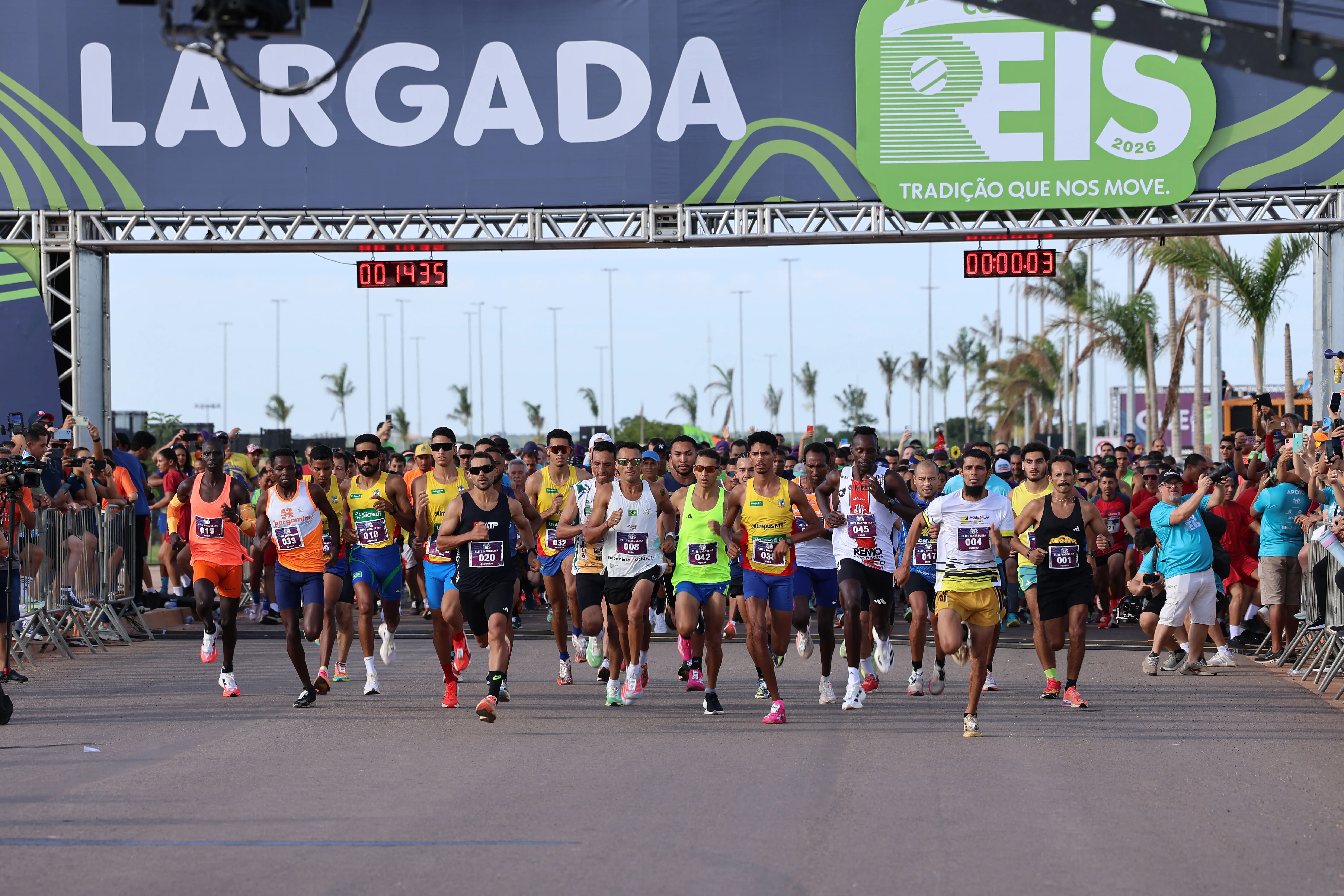 Atletas largando na Corrida de Reis 2026. Corredores de diversos perfis passam sob o portal oficial de largada em uma pista de asfalto sob céu claro.
