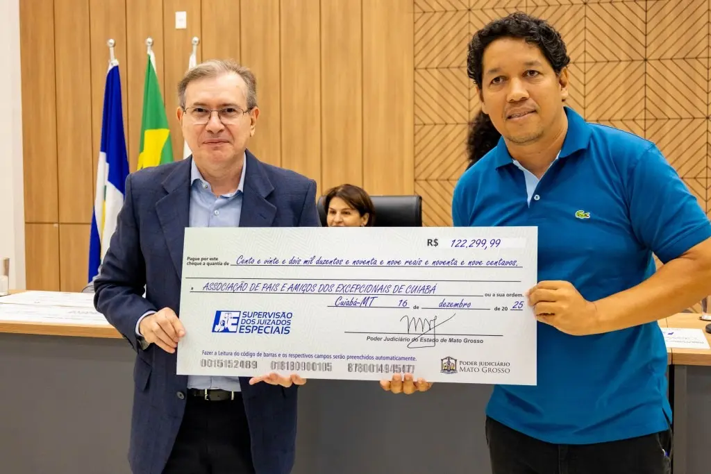 Desembargador entrega cheque simbólico a representante da Apae   durante cerimônia no auditório do Complexo dos Juizados. Eles estão em pé, em frente à mesa de autoridades.