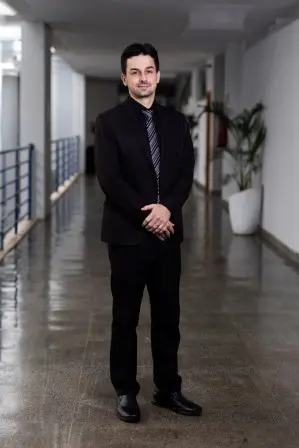 Foto do diretor do DAPI. Homem de pele clara, cabelo curto escuro. Veste terno preto, camisa preta e gravata preta com listras discretas. Está com as mãos unidas à frente do corpo e olha para a câmera