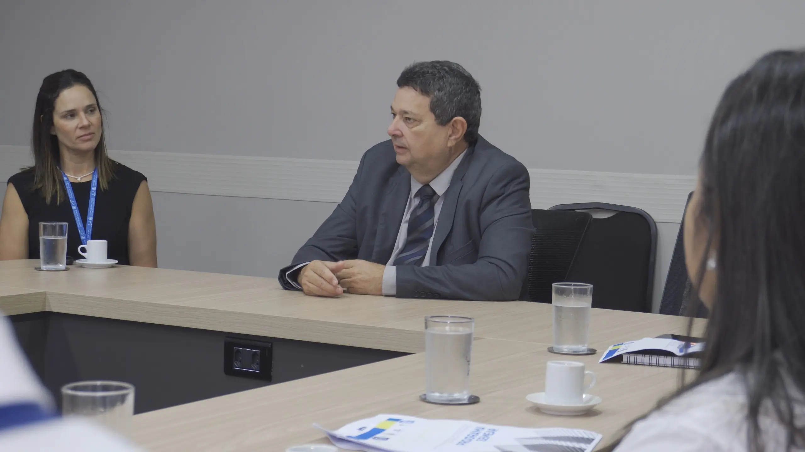 Homem de terno cinza e gravata fala em uma mesa de reunião clara. Ao fundo, uma mulher observa. Copos de água e xícaras de café estão sobre a mesa.