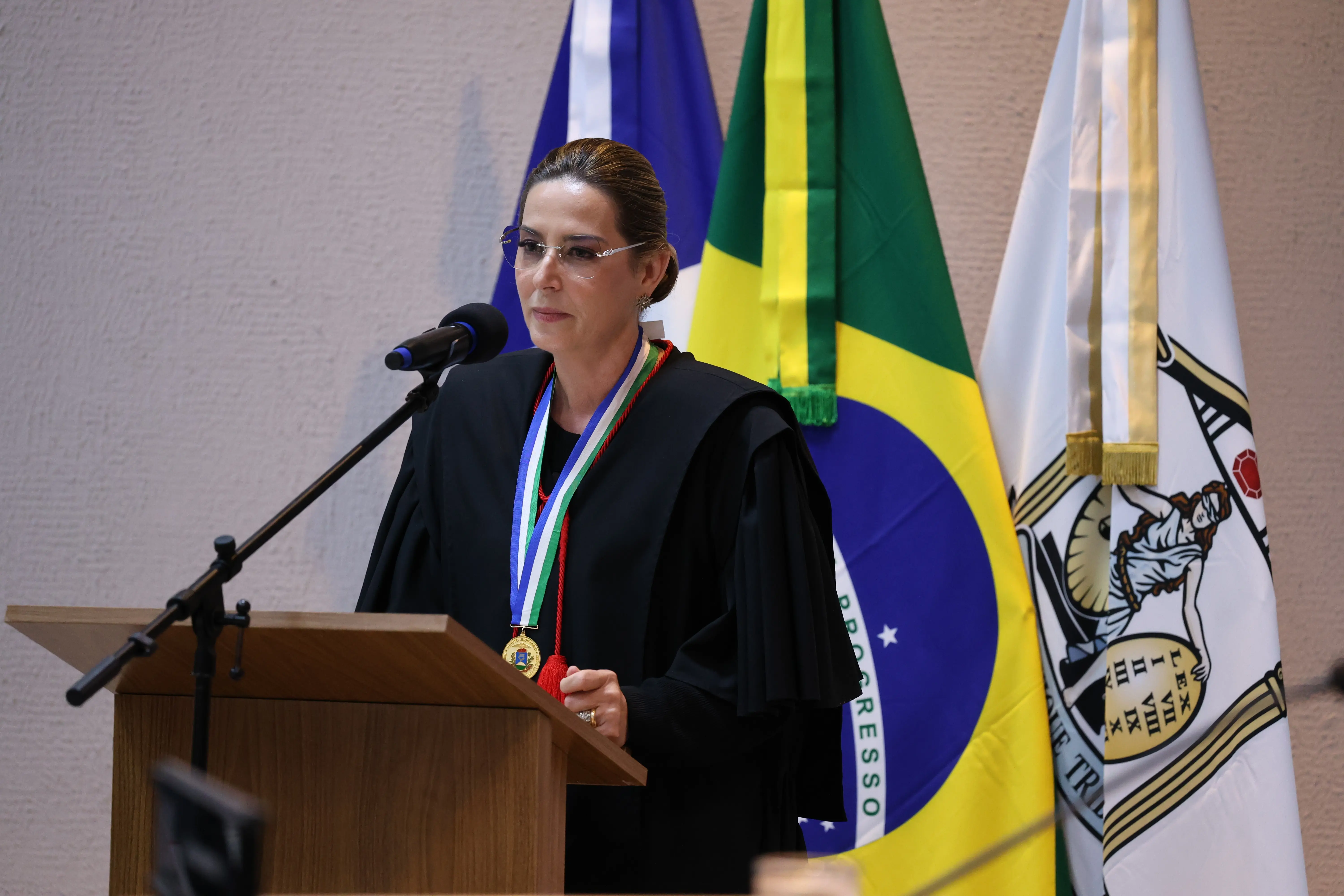 Mulher discursa ao microfone em púlpito, vestindo traje de magistrado e medalha tricolor. Ao fundo, bandeiras do Brasil e de Mato Grosso, além de brasão do TJMT.