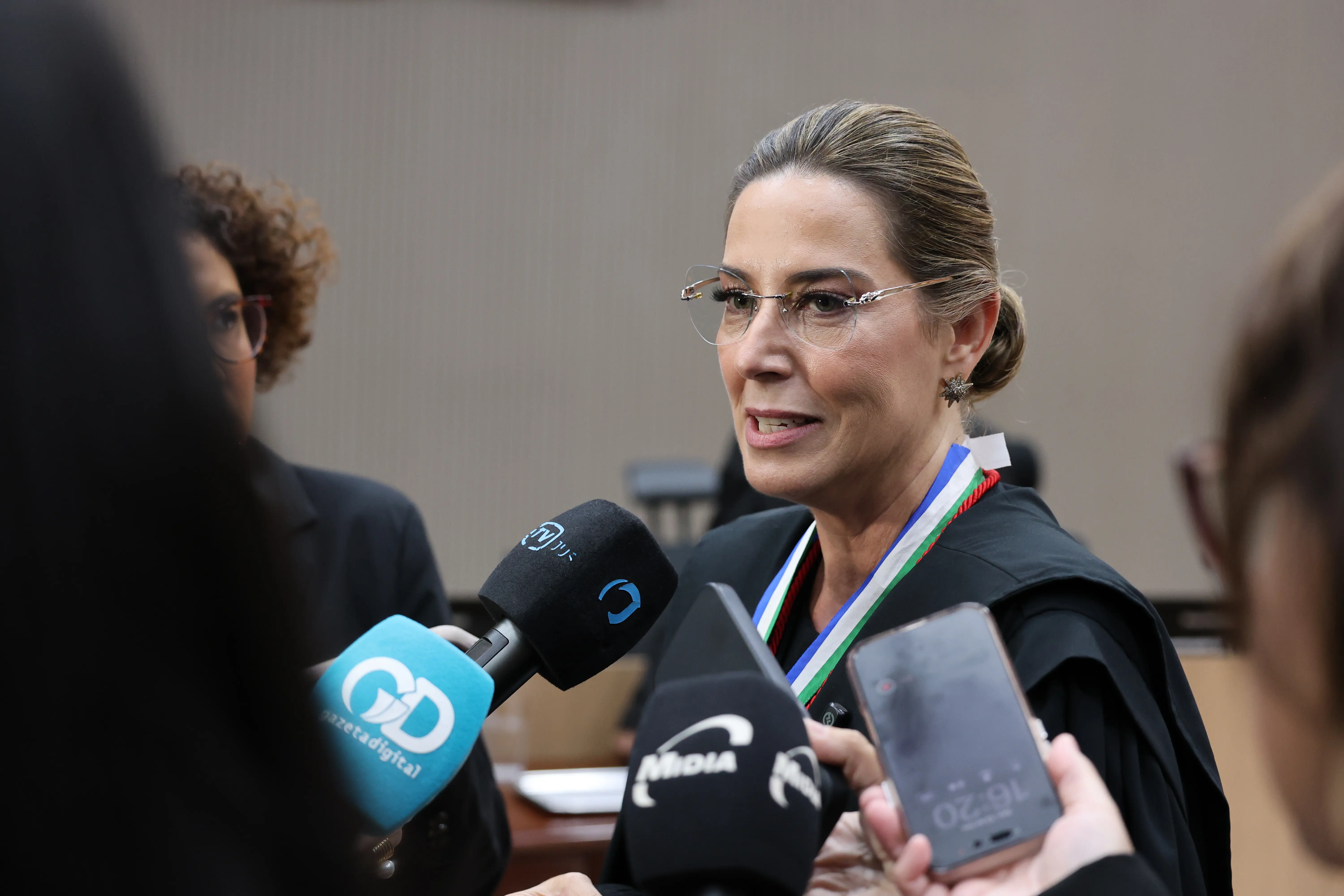 Mulher de traje de magistrado e medalha tricolor concede entrevista cercada por microfones de emissoras de TV. Ao fundo, pessoas e ambiente de tribunal. 