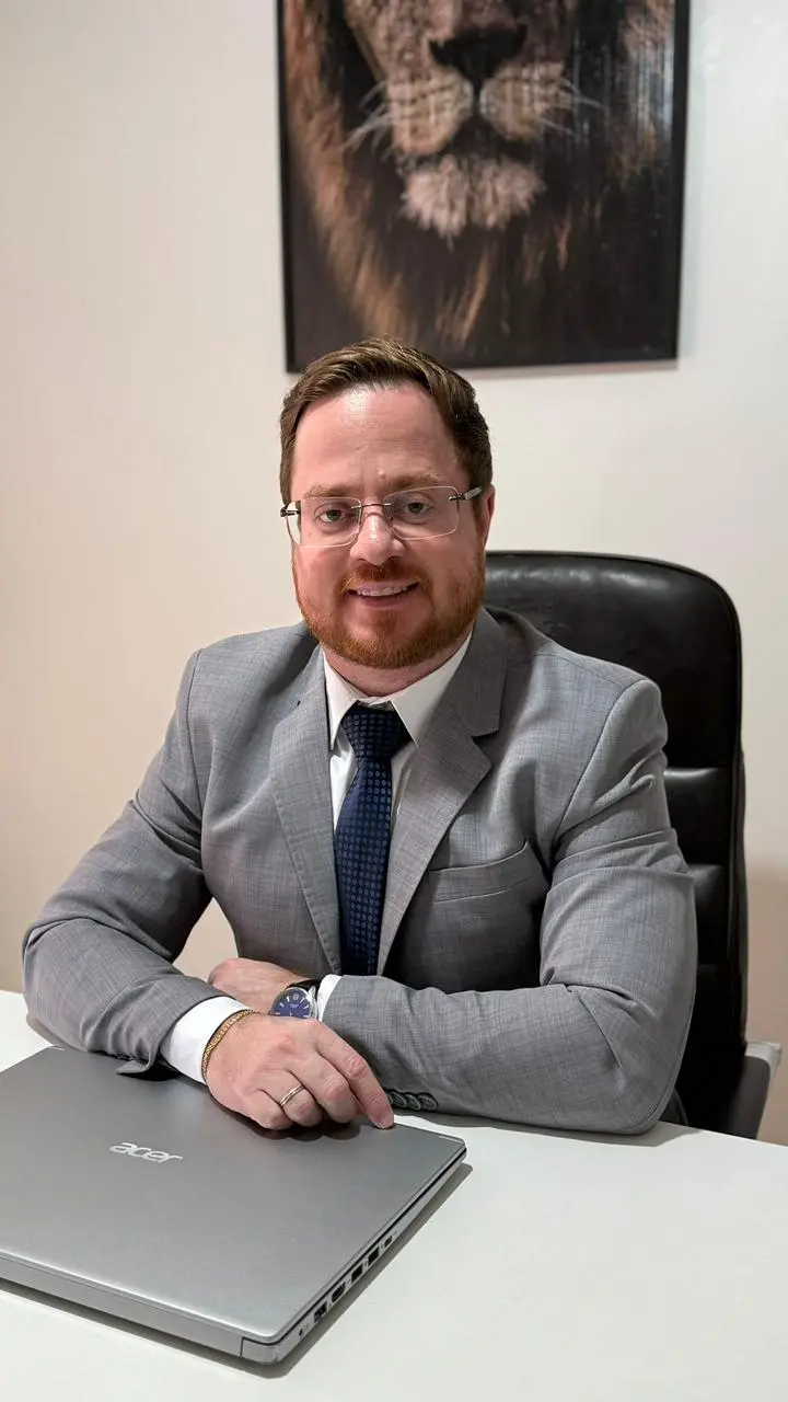 Foto do advogado. Homem de pele clara, cabelo curto castanho e barba ruiva, sentado em cadeira preta atrás de mesa de escritório. Usa óculos, terno cinza, camisa branca e gravata azul. Apoia os braços sobre a mesa, onde há um notebook fechado. Ao fundo, parede clara com quadro de um leão