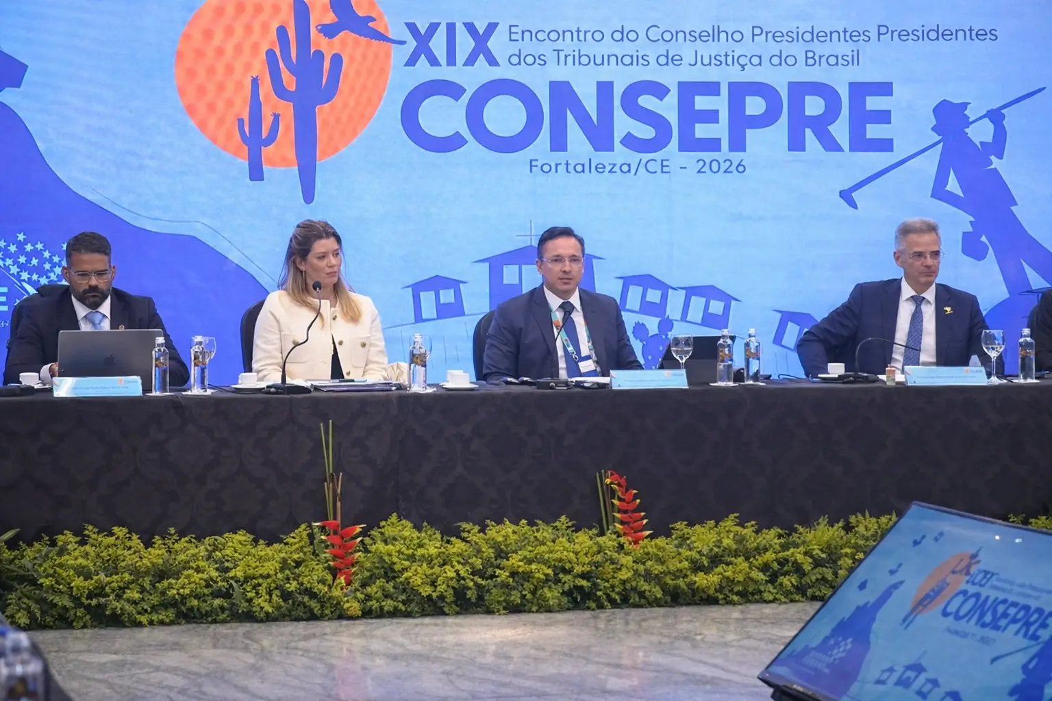 Mesa de autoridades no XIX Consepre, em Fortaleza, com magistrados sentados atrás de bancada preta, microfones e águas. Ao fundo, painel azul com logomarca do evento e elementos gráficos regionais.