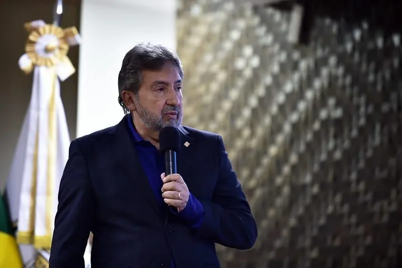 Homem de cabelo e barba grisalhos fala ao microfone. Veste paletó escuro sobre camisa azul. Ao fundo, à esquerda, há uma bandeira e, à direita, parede com painel decorativo.