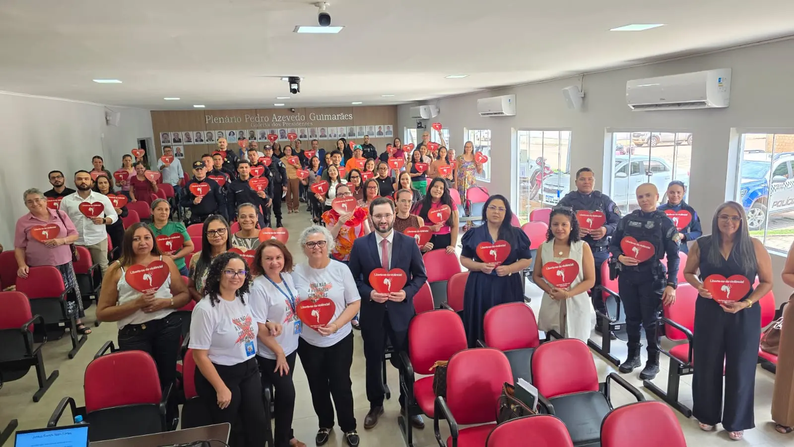 Foto em plano aberto que mostra dezenas de pessoas no plenário da Câmara Municipal de Porto Alegre do Norte. Elas sorriem para a foto e seguram panfletos vermelhos em formato de coração.