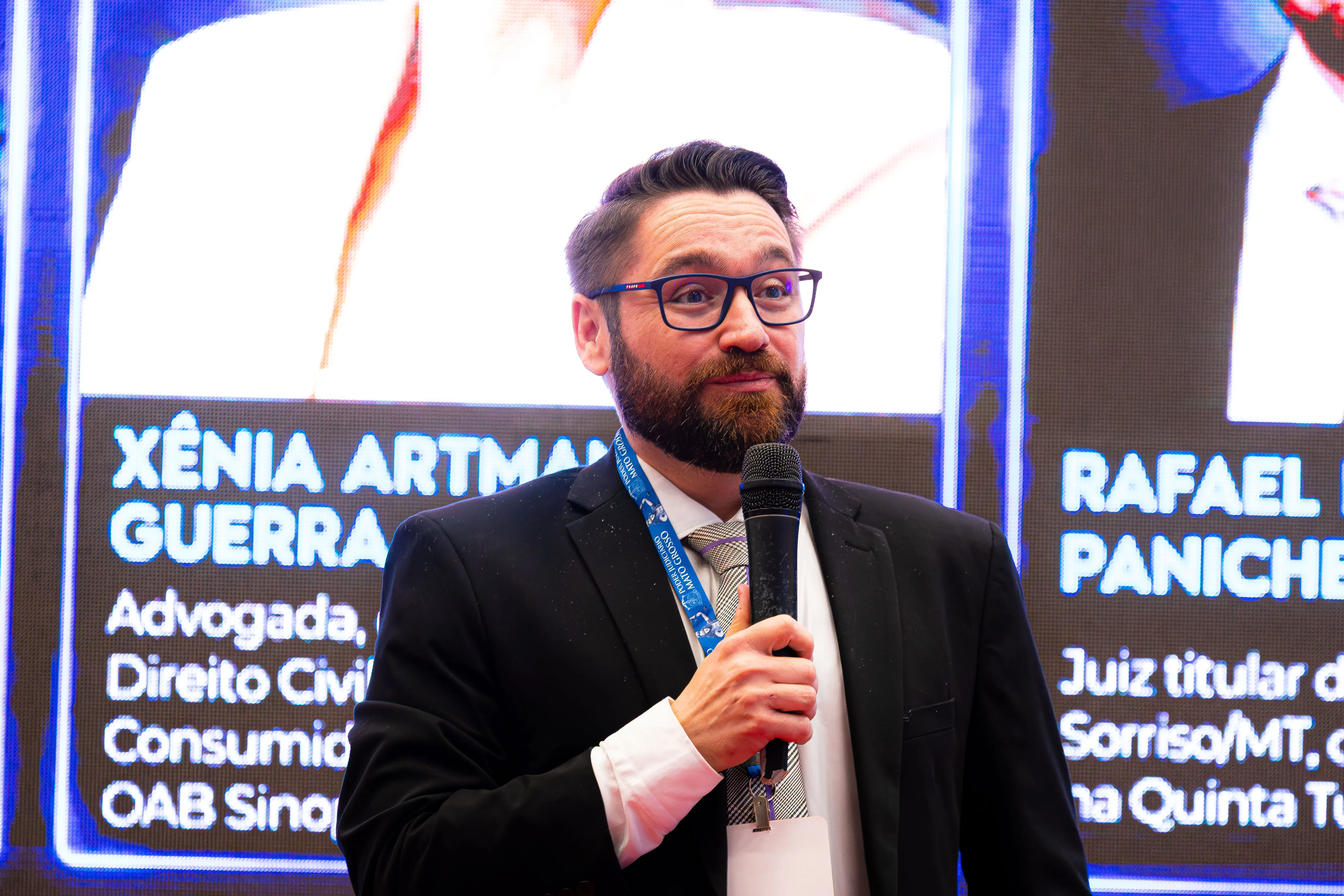 Homem de óculos, barba e terno preto fala ao microfone. Ele usa crachá com cordão azul. Ao fundo, um painel luminoso exibe fotos e textos sobre palestrantes.