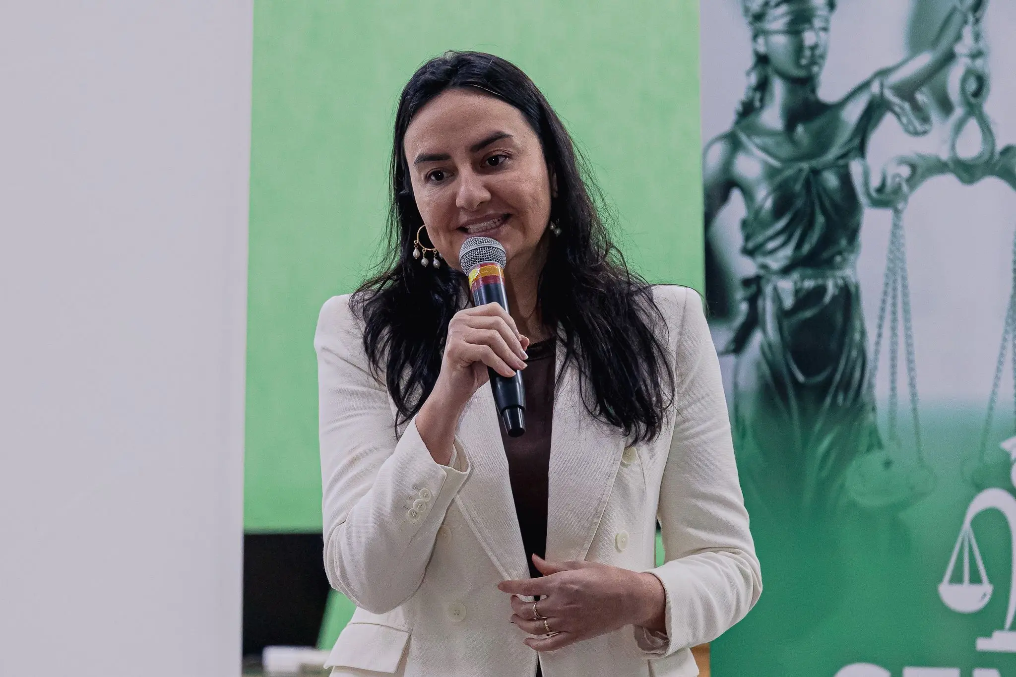 Foto de mulher de cabelos pretos e longos, vestindo um paletó branco sobre uma blusa escura, que segura um microfone com a mão direita próxima à boca enquanto fala. Ao fundo, painel desfocado em tom verde-claro.