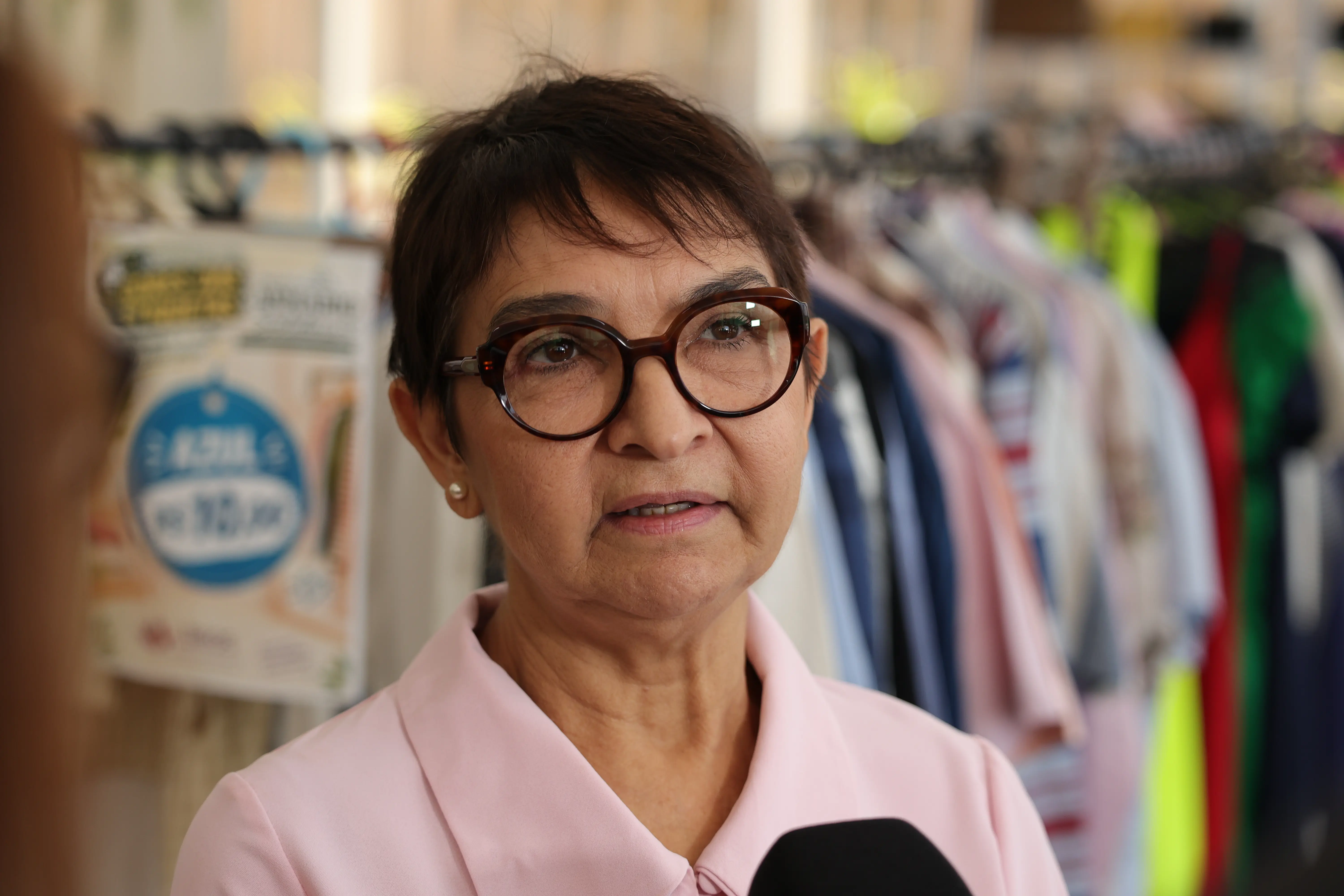 close-up de uma mulher com óculos de aro marrom e brinco de pérola. Ela veste uma blusa rosa claro. O fundo está embaçado e mostra araras de roupas e um microfone na parte inferior.