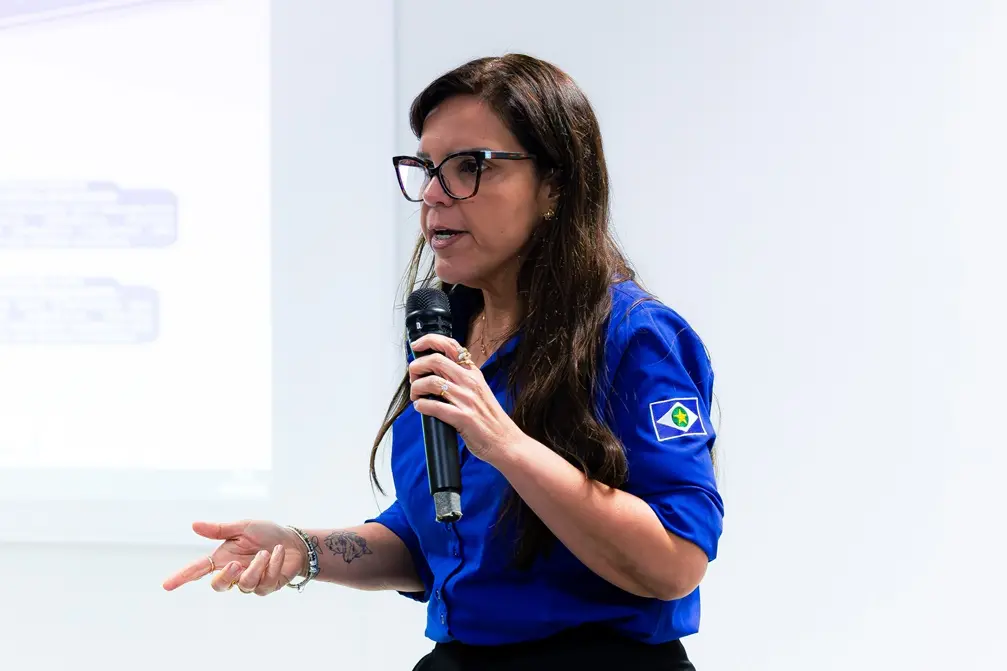 Uma mulher de cabelos longos castanhos e óculos de armação escura segura um microfone com a mão direita e gesticula com a esquerda. Ela veste uma camisa azul com o brasão estampado na manga e possui uma tatuagem visível no antebraço esquerdo.