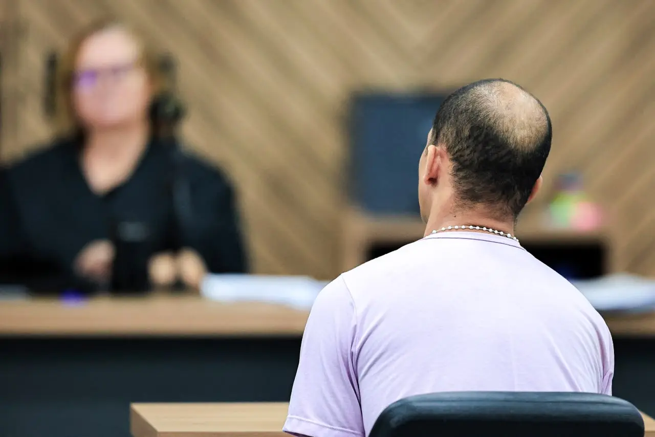 Foto horizontal que mostra um homem branco e calvo, sentado no banco do réu, diante da juíza, que aparece desfocada na foto. O homem está de costas para a foto, ele usa camiseta lilás.