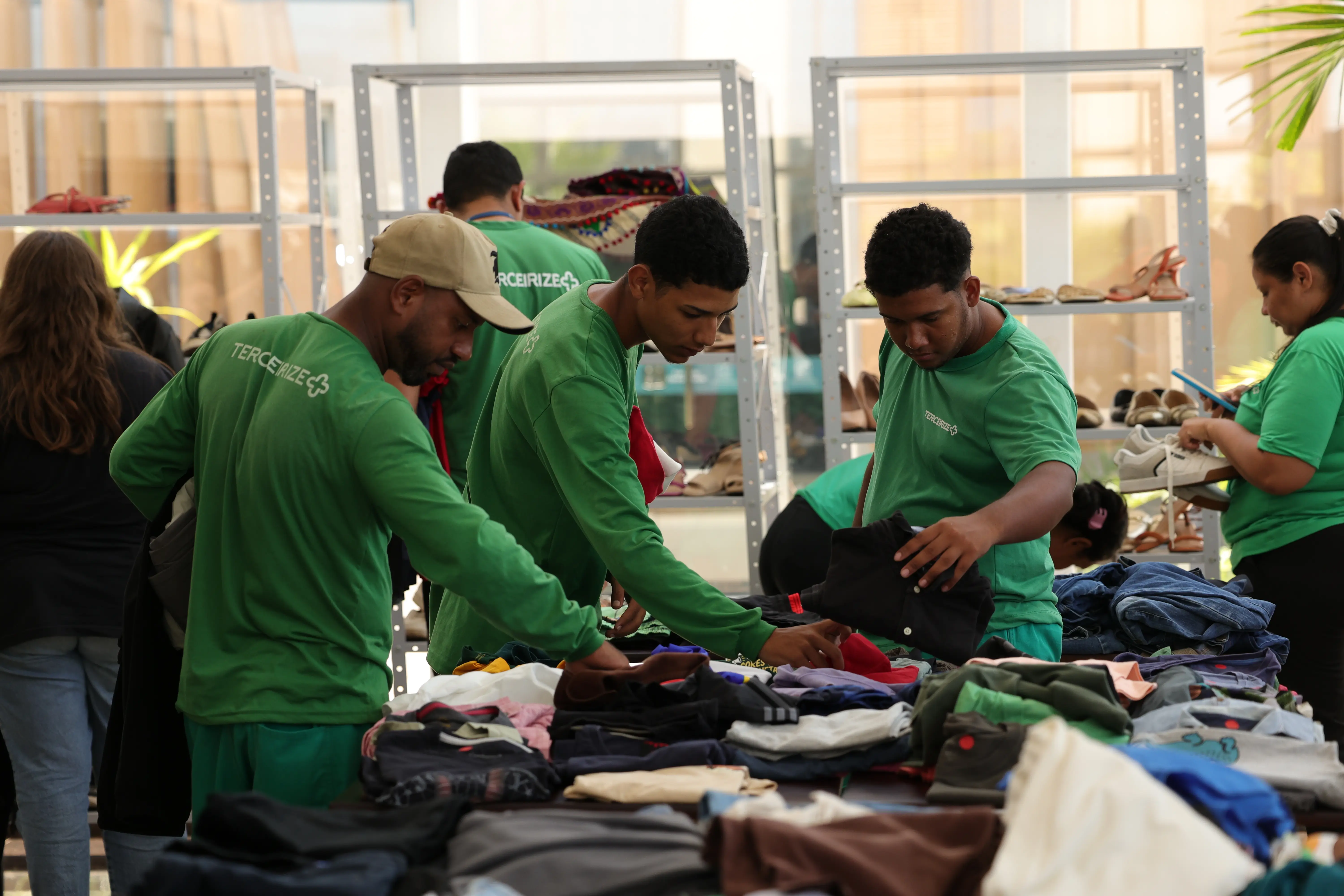 Homens vestindo camisetas verdes de manga longa e boné manuseiam pilhas de roupas dobradas em mesas. Uma mulher de camiseta verde está ao fundo à direita, também mexendo em roupas em prateleiras de metal cinza. O ambiente é amplo e claro.