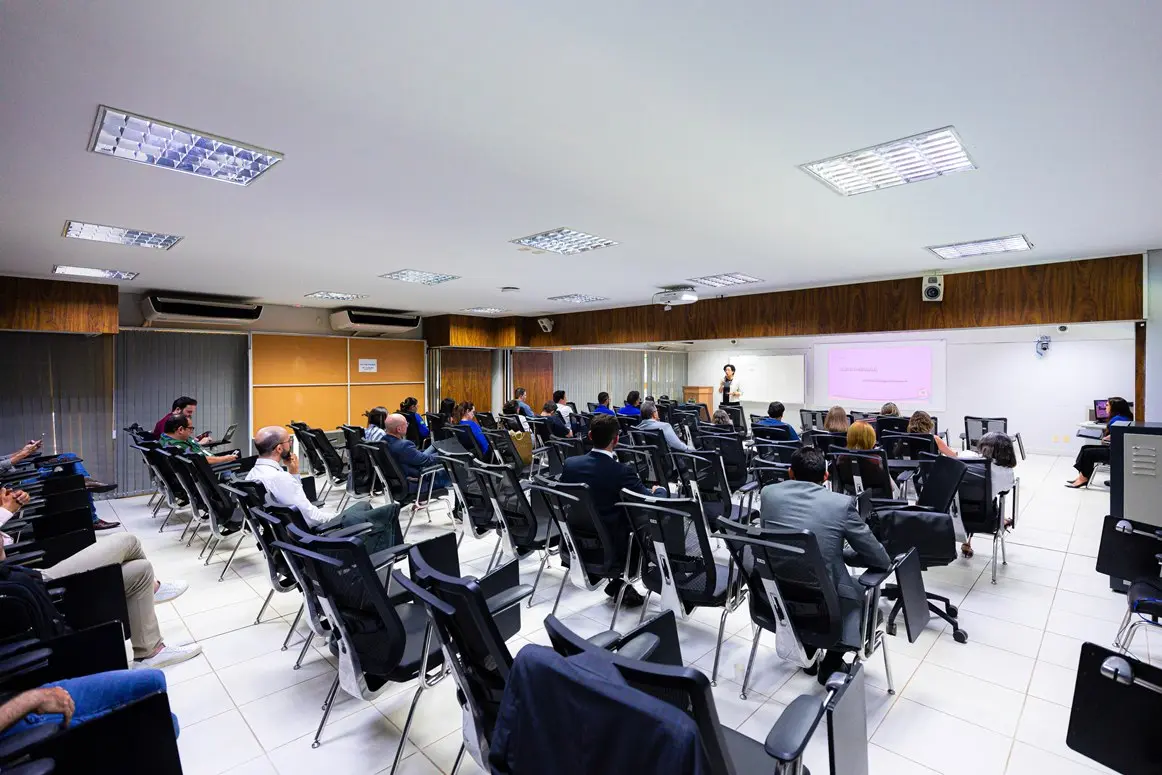 Vista ampla de um auditório iluminado com teto claro e painéis de madeira ao fundo, onde várias pessoas assistem a uma apresentação. Na frente, uma palestrante em pé fala ao lado de uma tela de projeção e um quadro branco, diante de várias fileiras de cadeiras pretas ocupadas.