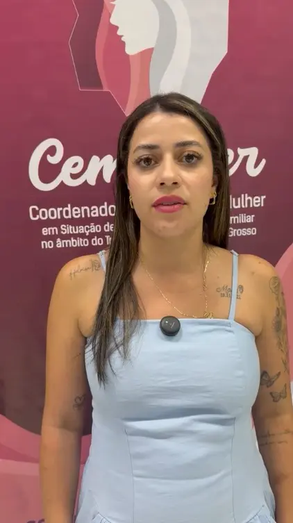Foto vertical que mostra a advogada Letícia Gomes, da cintura pra cima, em pé em frente a uma banner cor-de-rosa da Cemulher. Ela é uma mulher jovem, de pele clara, cabelo castanho com mechas loiras, liso e comprido, usando vestido azul claro de alcinha.
