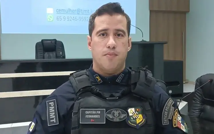 Foto horizontal que mostra o capitão PM Jeorge Fernandes, do busto pra cima, usando farda azul marinho da PM de Mato Grosso. Ele é um homem branco, de olhos e cabelos castanhos. Ele está em um auditório.