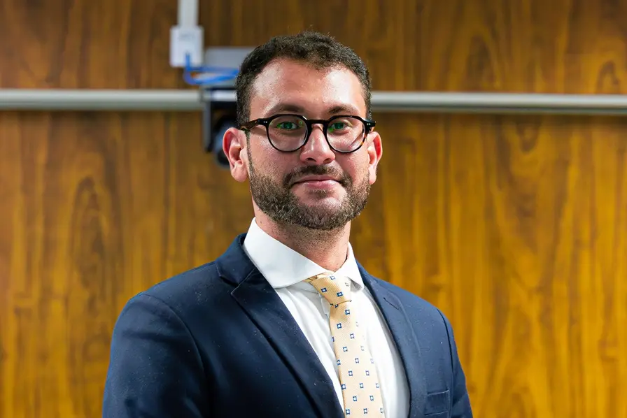 Homem de cabelos curtos e escuros, barba aparada e óculos de armação redonda preta, sorrindo levemente em enquadramento de primeiro plano. Ele veste paletó azul-marinho, camisa social branca e uma gravata amarela com pequenos detalhes geométricos escuros.
